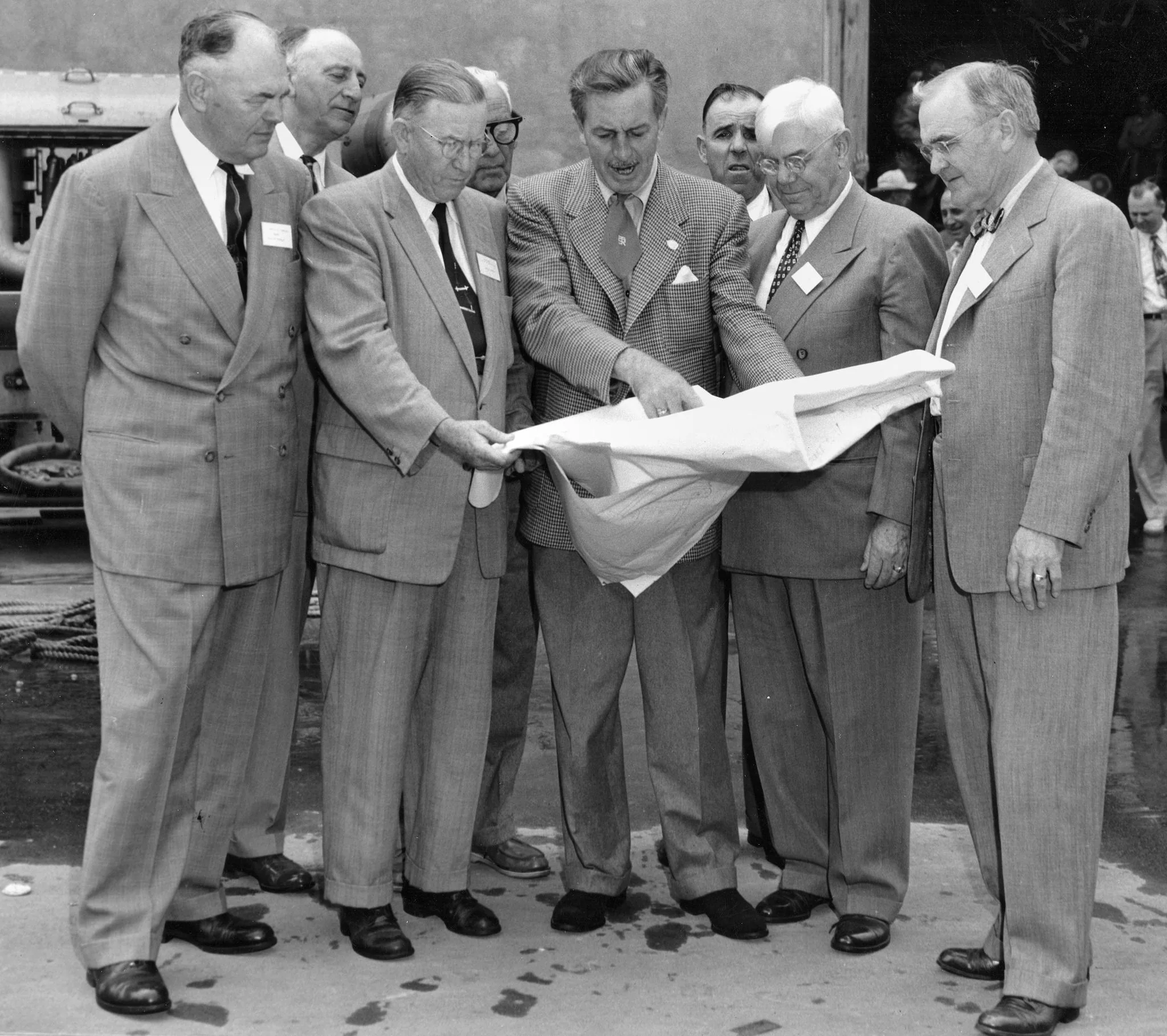 Walt Disney is depicted in this black-and-white photograph, surrounded by a group of well-dressed men, likely engaged in a planning or discussion session. The men are all wearing suits, showcasing the formal nature of the meeting. Walt Disney, positioned centrally, is actively gesturing towards a large sheet of paper, possibly a blueprint or plan. This suggests that he is leading or explaining a project. The group appears attentive, intently focused on the document in Walt's hands.
The setting seems to be outdoors, possibly at a construction site or an area under development. The background is indistinct but hints at industrial elements, like machinery or a construction area, reinforcing the context of planning or development. The atmosphere is serious yet collaborative, with the men closely huddled together, indicating a shared focus on the task at hand. The image captures a historical moment, reflective of an era of significant development and innovation led by Walt Disney.