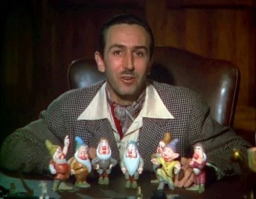 Walt Disney is seated at a desk, appearing younger, in an indoor setting. He is dressed in a checkered suit jacket with a white shirt and a patterned scarf, exuding a mid-20th-century style. In front of him on the desk, there are figurines of the seven dwarfs from the classic animated film "Snow White and the Seven Dwarfs." These figurines are arranged neatly, showcasing characters like Sleepy, Grumpy, Doc, and others, each with distinctive facial expressions and colorful outfits. The background features wooden paneling, adding a warm and cozy atmosphere to the scene. Walt Disney looks directly at the viewer, with a slight smile, embodying a sense of pride and enthusiasm, likely discussing or presenting his work. The lighting is soft, highlighting both Disney and the figurines prominently, giving the image an inviting and nostalgic feel.