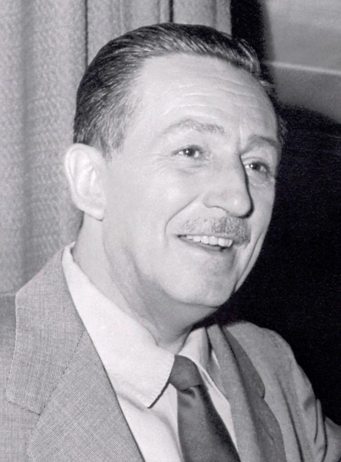 The image is a black-and-white portrait of Walt Disney. He is smiling warmly, creating an inviting and approachable aura. He is dressed in a formal suit with a tie, reflecting a mid-20th-century style. His hair is neatly combed back, and he sports a thin mustache, a characteristic look he was known for. The background is simple, possibly a curtain or a plain wall, emphasizing the focus on Walt Disney himself. The lighting is soft, highlighting his facial features gently, indicating that this might be a studio or professional setting. The monochromatic tones add a timeless quality to the portrait, capturing Walt Disney’s iconic presence in a way that feels both historical and enduring.