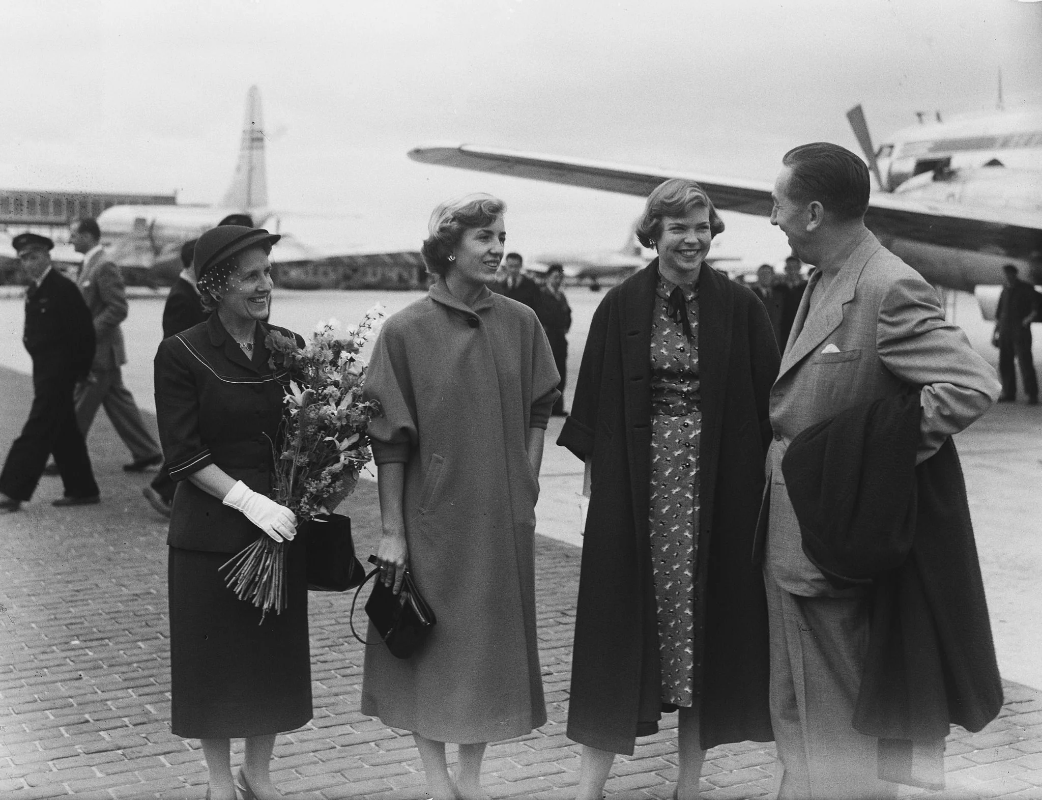 In the black-and-white photograph, Walt Disney is seen at an airport, engaging in conversation with three women. The setting is outdoors, with an airplane visible in the background, suggesting a scene of arrival or departure. Walt Disney, dressed in a suit, appears relaxed and engaged, holding a coat over his arm. The woman on the left is holding a bouquet of flowers, possibly as a welcoming gesture. She is dressed in a coordinated suit and hat, wearing gloves, indicative of the fashion of the time. The other two women are also dressed stylishly, one in a patterned dress with a coat draped over her shoulders and the other in a simple, elegant coat. They are all smiling, creating a warm and sociable atmosphere. The background shows more people and another airplane, emphasizing the busy and bustling nature of the airport. The photograph captures a moment of casual interaction and hospitality.