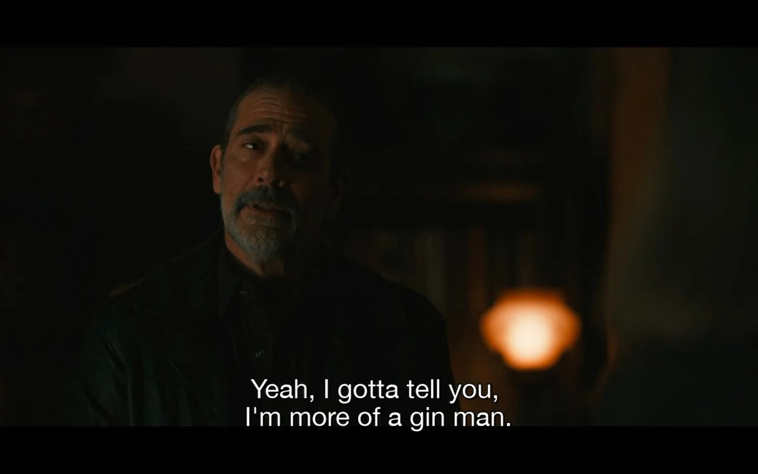 Jeffrey Dean Morgan is depicted in this scene from "TWD," portraying his character against a dimly lit background, suggesting an intimate or tense atmosphere. He appears to be engaged in a conversation, with a focused expression that conveys sincerity or conviction. The lighting casts shadows across his face, enhancing the dramatic effect of the moment. He is dressed in a dark jacket, which blends into the shadowy surroundings, providing a somber tone. The scene is likely set indoors, as suggested by the blurred background elements, including what appears to be shelves or furniture. The subtitle reads, "Yeah, I gotta tell you, I'm more of a gin man," indicating a personal revelation or a moment of levity within the dialogue. This setting and mood suggest a scene that combines elements of drama with a touch of humor, characteristic of the nuanced character interactions in the show.
