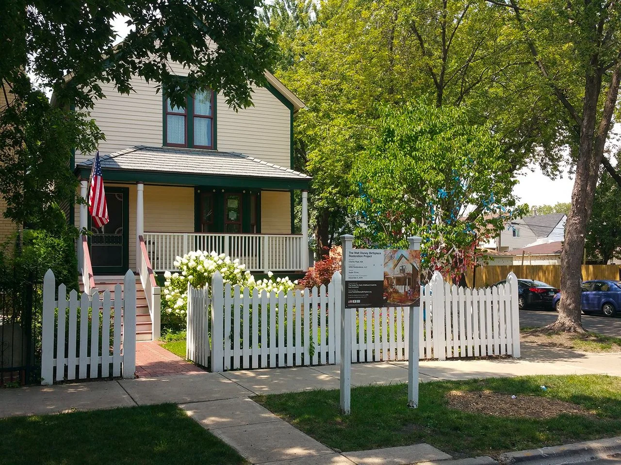 The image depicts a quaint, historical house known as "The Blue House," which is the birthplace of Ernest Hemingway. The house is a two-story structure with beige siding and green trim, giving it a classic and inviting appearance. An American flag is mounted near the entrance, highlighting its national significance. The front yard is enclosed by a charming white picket fence, with a gate opening onto a brick path leading to a small porch. The porch is adorned with white railings and a pair of white columns.
In the foreground, lush greenery and blooming white flowers add to the picturesque setting. A sign in the yard reads "The Blue House Birthplace of Ernest Hemingway," providing context to visitors. Large trees in the background offer shade, and a sidewalk runs in front of the house. The scene is set on a sunny day, evidenced by the bright lighting and clear sky, contributing to the warm and welcoming atmosphere of this historical landmark.