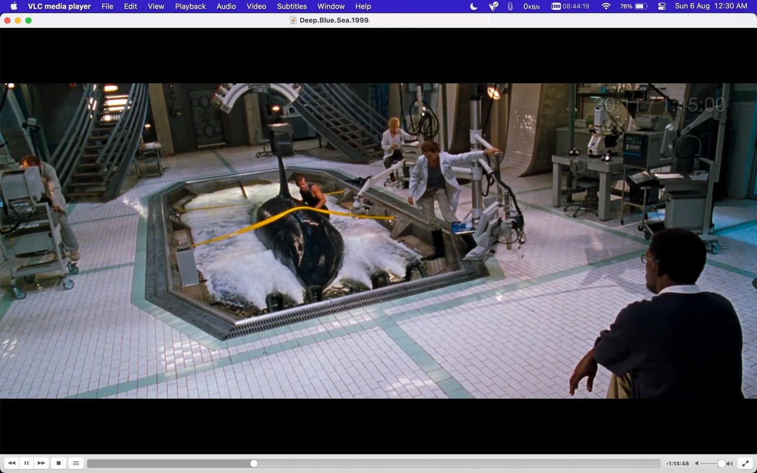 The image is a frame from the film "Deep Blue Sea," displayed on the VLC media player interface. The scene takes place in a high-tech laboratory setting. In the center, a large shark is partially submerged in a water tank, surrounded by lab equipment and several scientists engaged in an experiment. One scientist, dressed in a white lab coat, is operating machinery, while another is interacting closely with the shark. The setting is well-lit, with tiled floors and industrial details like metal staircases and control panels in the background. The atmosphere is tense and focused, reflecting the scientific exploration theme. The VLC media player's interface is visible along the top and bottom of the frame, showing the video file name and playback controls. The title bar reads "Deep Blue Sea 1999 1080p B...," indicating the movie being viewed.