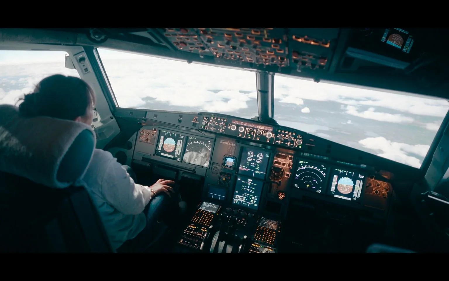 The image depicts the interior of an airplane cockpit, likely during a flight. A pilot is seated on the left side, wearing a light-colored shirt and appearing focused on the task of flying the aircraft. The cockpit is filled with an array of controls, switches, and screens that display various flight data, such as altitudes and navigational information. The atmosphere is technical and professional, with a mix of digital and analog instruments that are typical in modern aircraft.
Outside the cockpit windows, the view shows a bright sky dotted with clouds, suggesting the plane is at cruising altitude. The lighting inside the cockpit is subdued, with the glow of the instruments providing a contrast to the bright daylight outside. The image captures the complexity and precision required in aviation, as well as the advanced technology pilots rely on to safely navigate the skies.