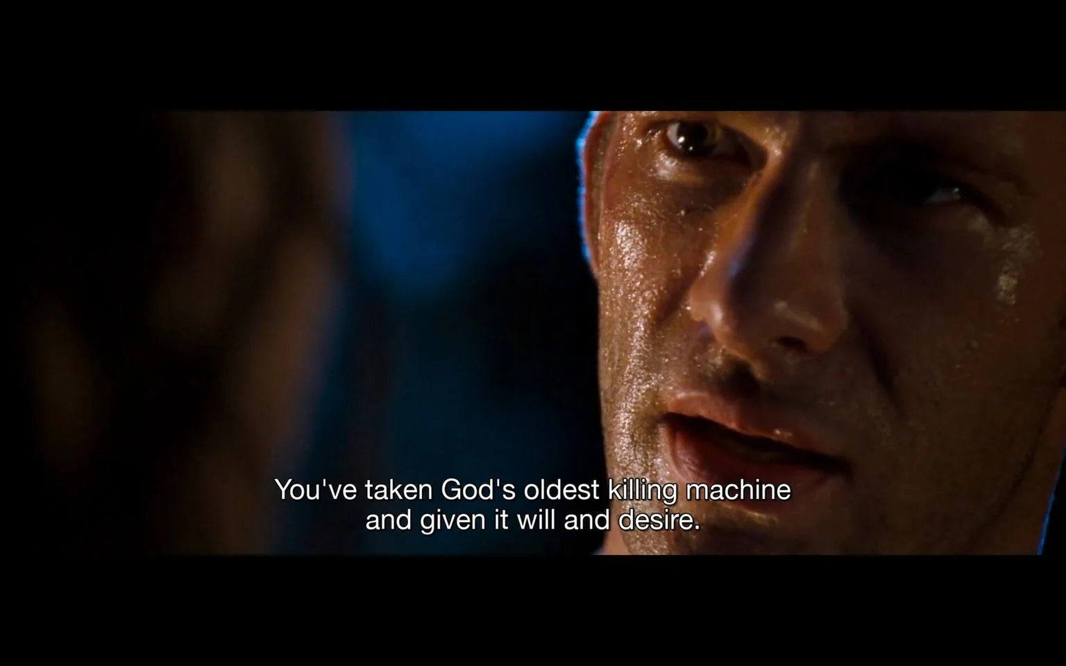 The image is a dramatic close-up of a man's face, captured during an intense moment in a film. His face is glistening, suggesting a high-stakes or tense situation, possibly under harsh lighting conditions. The background is blurred and dark, which enhances the focus on his expression—serious and contemplative. The lighting casts shadows that highlight the contours of his face, adding to the intensity of the scene.
The subtitle at the bottom reads, "You've taken God's oldest killing machine and given it will and desire," hinting at a conversation about a powerful and potentially dangerous entity. The mood conveyed is one of urgency and gravity, underscoring a critical or pivotal moment in the narrative. The use of language suggests themes of creation, control, and perhaps ethical dilemmas. The scene is likely set at night, with a somewhat somber and serious tone, characteristic of suspenseful or thriller genres.