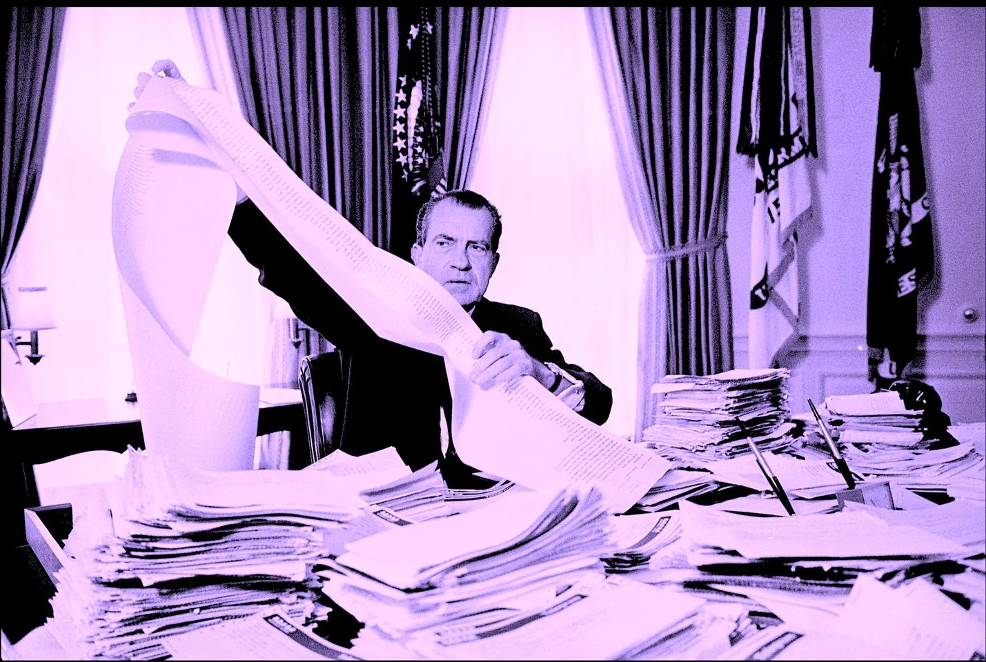 The image features Richard Nixon, as indicated by the hint, seated at a large office desk covered with numerous stacks of papers. He is in the process of examining a long, continuous sheet of paper, which he holds up for review. The setting appears to be an official office, likely the Oval Office, given the grandeur of the surroundings. Behind him, large draped curtains frame the windows, and flags stand on either side, enhancing the formal atmosphere.
Nixon is wearing a dark suit, typical of his presidential attire, and his expression conveys concentration and seriousness as he reviews the documents. The lighting casts a purplish tint over the image, giving it a somewhat vintage feel. The desk is cluttered with papers, folders, and pens, suggesting an environment of intense work and documentation. This scene captures a moment of administrative or decision-making processes during Nixon's presidency.
