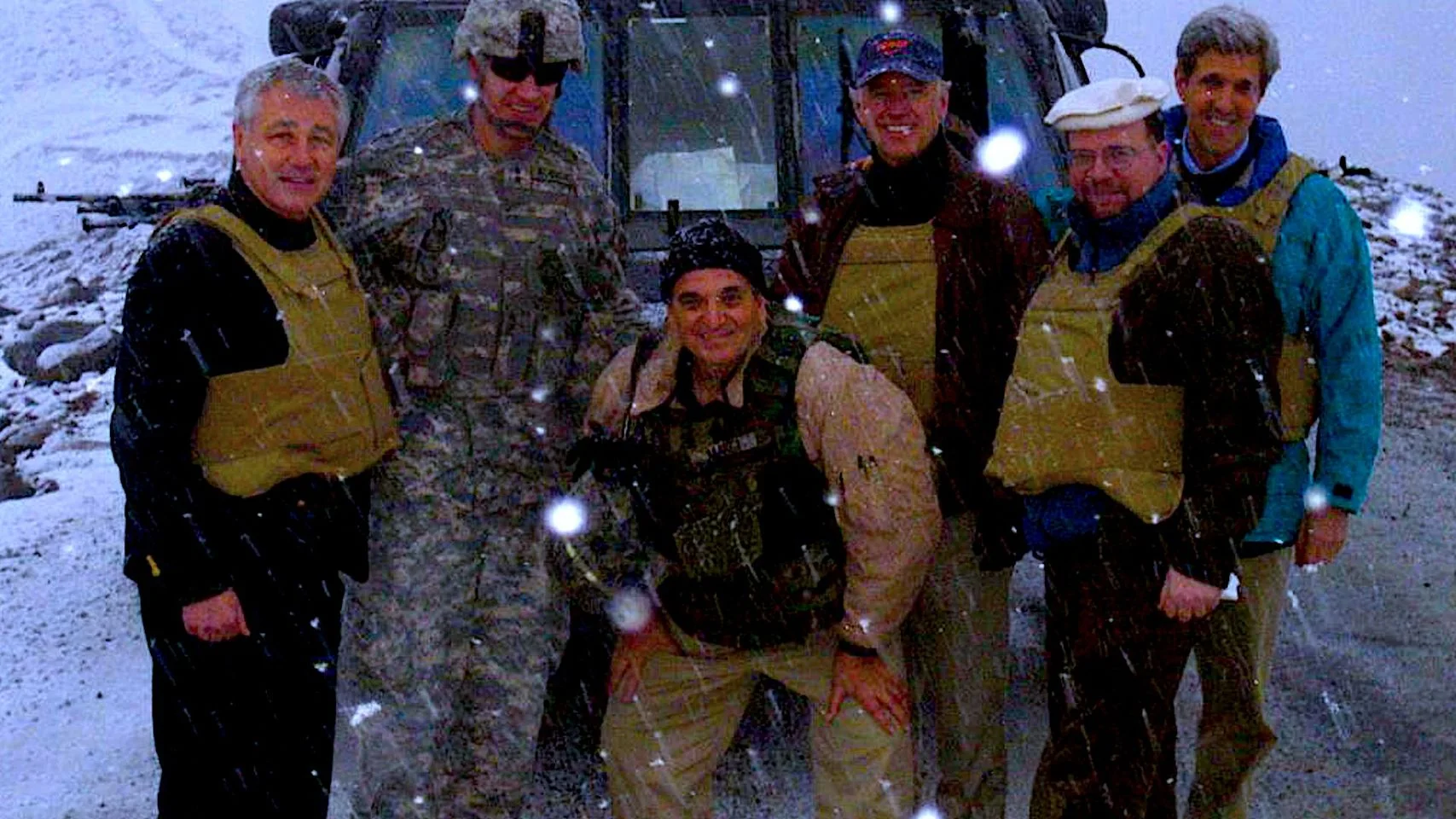 In the image, a group of men is standing in a snowy outdoor setting. They are dressed in a mix of military and civilian attire, with several wearing protective vests. The central figure in camouflage appears to be a soldier, suggesting a military setting. The hint provided identifies one of the individuals as John Kerry, who is standing on the right side of the group. The men are positioned in front of a vehicle, possibly a military transport, partially visible behind them. The mood seems amiable and cooperative, as they are smiling despite the snowfall. The lighting is natural, and the snowflakes are visibly captured in the frame, adding to the chilly atmosphere of the scene. The overall composition is casual, indicative of a moment captured during a visit or inspection in a cold, possibly mountainous region.