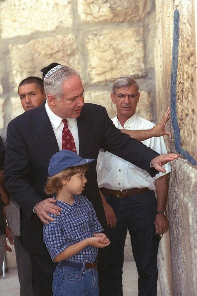 ```
Benyamin Netanyahu is seen in this image, engaging with a young boy at a historical site, likely in Jerusalem, given the stone walls and significant cultural context. Netanyahu, wearing a dark suit and a red patterned tie, also has a kippah on his head, indicating the religious significance of the location. He is attentively gesturing towards the wall, possibly explaining something of importance. The young boy, dressed in a blue checkered shirt and blue jeans, is wearing a baseball cap and seems to be listening intently. Around them, a few other men are present, one of whom is touching the wall with a contemplative expression. The setting is outdoors, under natural daylight, and the mood appears educational and respectful. The composition highlights Netanyahu's role as a guide or mentor, emphasizing cultural and historical appreciation.