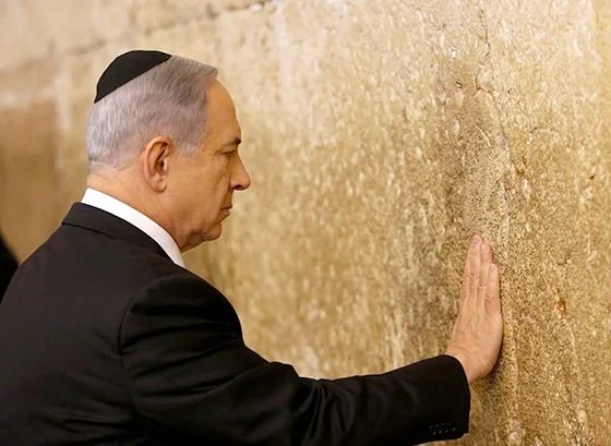 Benyamin Netanyahu is depicted in this image, standing at the Western Wall, a significant historical and religious site in Jerusalem. The photograph captures a solemn moment as he places his hand on the ancient stones, a gesture often associated with prayer or reflection. Netanyahu is dressed formally in a dark suit and white shirt, with a small black kippah on his head, signifying his respect for the religious customs of the location. The wall itself is textured with large, weathered stones, conveying a sense of history and reverence. The lighting is bright, suggesting daytime, and highlights the details of both the stone and Netanyahu's attire. The composition focuses on the interaction between Netanyahu and the wall, emphasizing a personal, contemplative moment. The overall mood is one of solemnity and respect, capturing the significance of the site within a cultural and historical context.