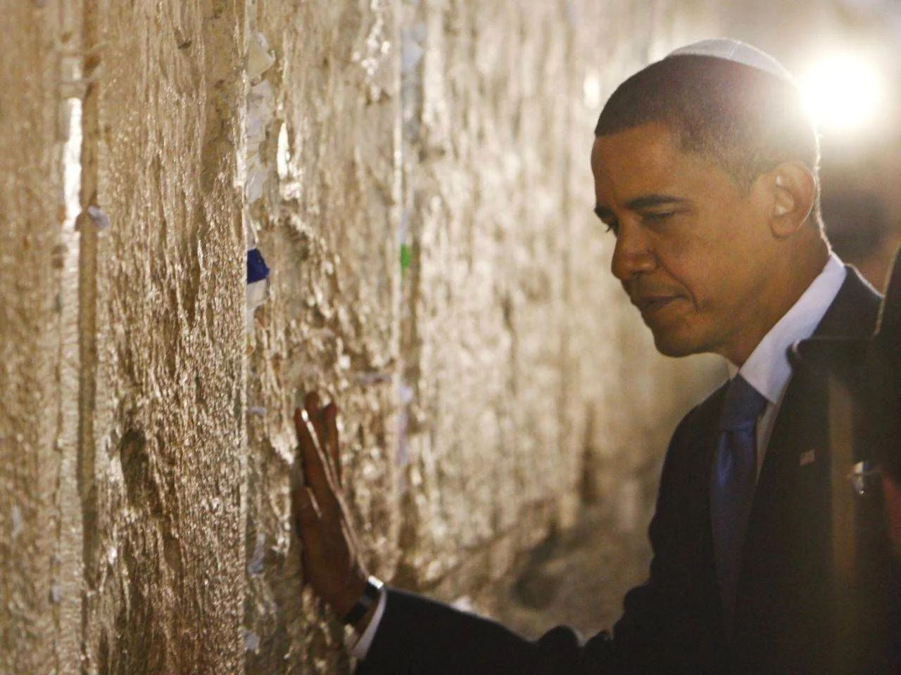 In this image, Barack Obama is shown at a historic site, placing his hand thoughtfully on the wall. He is wearing a dark suit with a light blue tie, and a kippah on his head, suggesting his participation in a moment of reflection or prayer. The stone wall is textured and has small pieces of paper inserted in its crevices, typical of the Western Wall, a place of significant cultural and religious importance. The lighting in the scene is soft, with a warm glow illuminating the area, creating a contemplative atmosphere. Obama's expression is one of contemplation and respect, enhancing the solemn mood of the setting. The background is softly blurred, focusing the viewer's attention on the interaction between Obama and the wall. The overall composition emphasizes the personal and reverent nature of the moment.