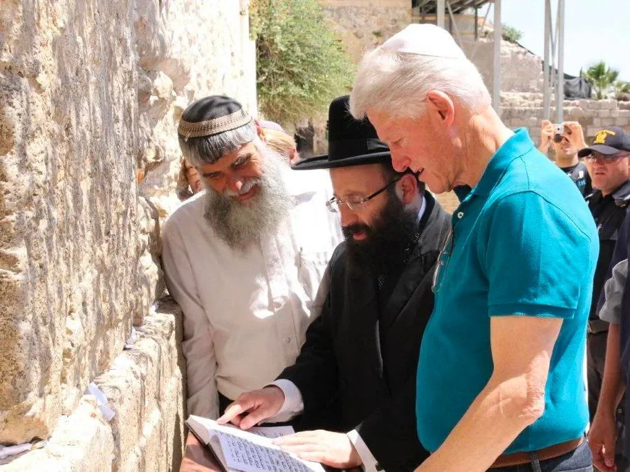 ```
1) Rich description: The image shows Bill Clinton, dressed casually in a blue polo shirt and wearing a kippah, standing at a historical site. He is accompanied by two men dressed in traditional Jewish attire, including one with a long white beard and another in a black hat and coat. The three are closely examining a book with Hebrew text, possibly engaging in a discussion or learning session. The setting is outdoors, likely at a significant cultural or religious landmark, as indicated by the stone wall beside them. Several people are visible in the background, some taking photos, suggesting that this is a public or semi-public event. The lighting is bright, indicative of a sunny day, which enhances the colors of the scene. The mood appears to be respectful and educational, with the individuals focused on the text in front of them.
2) On-screen text block:
- Title: "..."
- Channel / profile: "..."
- Site / app: "..."
- Captions / subtitles: "..."