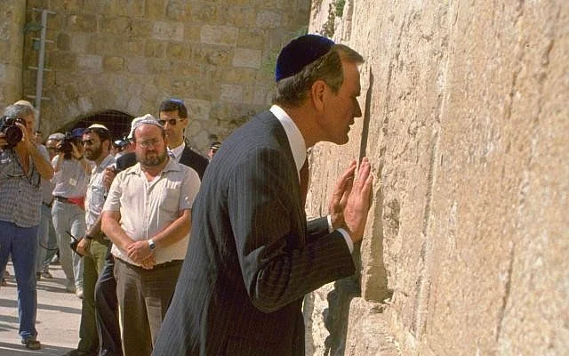 In the image, HW Bush is seen at the Western Wall, a significant historical and religious site. He leans forward with his hands touching the ancient stones, engaging in a moment of reflection or prayer. He is dressed in a formal dark suit and wears a blue kippah, which is customary for men at the site. The background shows the stone wall extending out of focus, with other individuals standing nearby, some observing and others capturing the moment on cameras. The lighting suggests a bright, sunny day, casting clear shadows and highlighting the texture of the stone. The scene is solemn and respectful, capturing a customary gesture of reflection at this sacred location. There is a sense of historical significance in the image, underscored by the presence of HW Bush and the iconic setting.