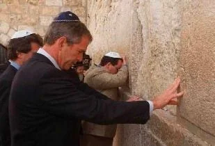 ```
The image depicts GW Bush at a significant historical and religious site, placing his hand on a large stone wall, which is likely part of a revered location such as the Western Wall in Jerusalem. He is accompanied by other individuals, all wearing traditional kippahs, signifying a moment of respect or prayer. GW Bush is wearing a formal dark suit, white shirt, and a dark tie, indicative of a formal visit or diplomatic event. The setting is outdoors, with the massive stone wall occupying most of the background, showcasing its historical texture and significance. The lighting suggests daytime, casting clear shadows and highlighting the solemn mood of the scene. The people present are engaged in what appears to be a solemn or reflective act, with one individual in the background leaning forward, possibly in prayer. The composition focuses on GW Bush in the foreground, emphasizing the importance of his presence and action in this context.