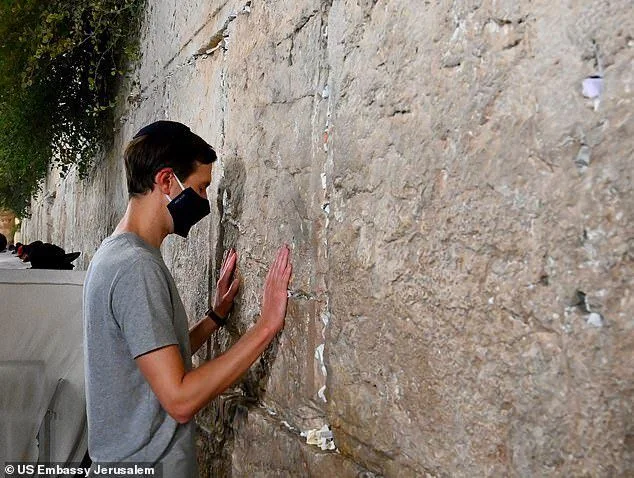 The image depicts a person standing in front of the Western Wall, a significant religious site in Jerusalem. The individual is engaged in prayer, with both hands placed on the ancient stone wall. They are wearing a black face mask and a gray t-shirt, adhering to health precautions and maintaining a respectful demeanor. The lighting is natural, suggesting the scene occurs during daylight hours. The wall is prominent, showcasing its worn, historic texture, and small pieces of paper, likely prayer notes, are visible tucked into its crevices. The background includes a partial view of more people, indicating the site's public nature. The image is credited to the US Embassy Jerusalem, highlighting its diplomatic context.