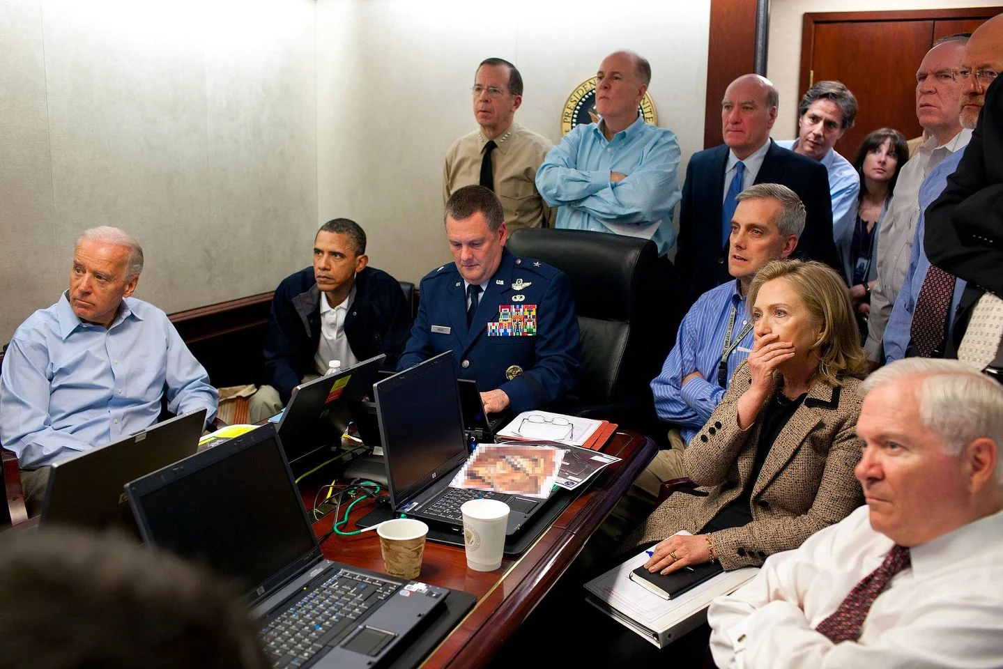 In this image, a group of officials are gathered in a conference room, focusing intently on something off-camera. Notable figures include Tony Blinken, who is standing towards the back. The setting appears to be a high-stakes meeting, indicated by the somber expressions and intense focus of those present. The room is filled with several laptops on the table, suggesting a briefing or monitoring of an ongoing situation. The participants are dressed in formal and semi-formal attire, with one military officer in uniform, adding to the serious and professional atmosphere. The lighting is bright and neutral, keeping the focus on the individuals and their expressions. Papers and disposable coffee cups are scattered around, hinting at a long or ongoing session. The overall mood is tense and concentrated, reflecting the gravity of the moment being captured. No specific on-screen text or titles are visible in the image.
