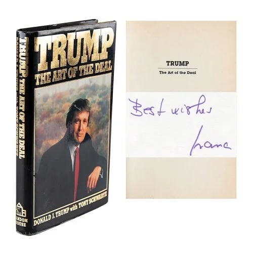 The image displays a book titled "Trump: The Art of the Deal." The book's cover prominently features the title in bold gold lettering with a photograph of a man in a suit, seated and looking confidently at the camera. The background of the cover includes a cloudy sky, adding to the dramatic effect of the presentation. The spine of the book, also visible, repeats the title and authors' names, emphasizing its branding.
Next to the book, there is a page from inside with text reading "TRUMP The Art of the Deal" at the top. Below this, there is a handwritten message: "Best wishes" followed by a signature. These elements suggest that the book is not only published by Random House but also personally signed, adding to its value and uniqueness. The overall composition focuses on the book and the autograph, highlighting its status as a collectible item. The dark tones of the cover are contrasted with the white and light colors of the inner page, creating a visual balance.