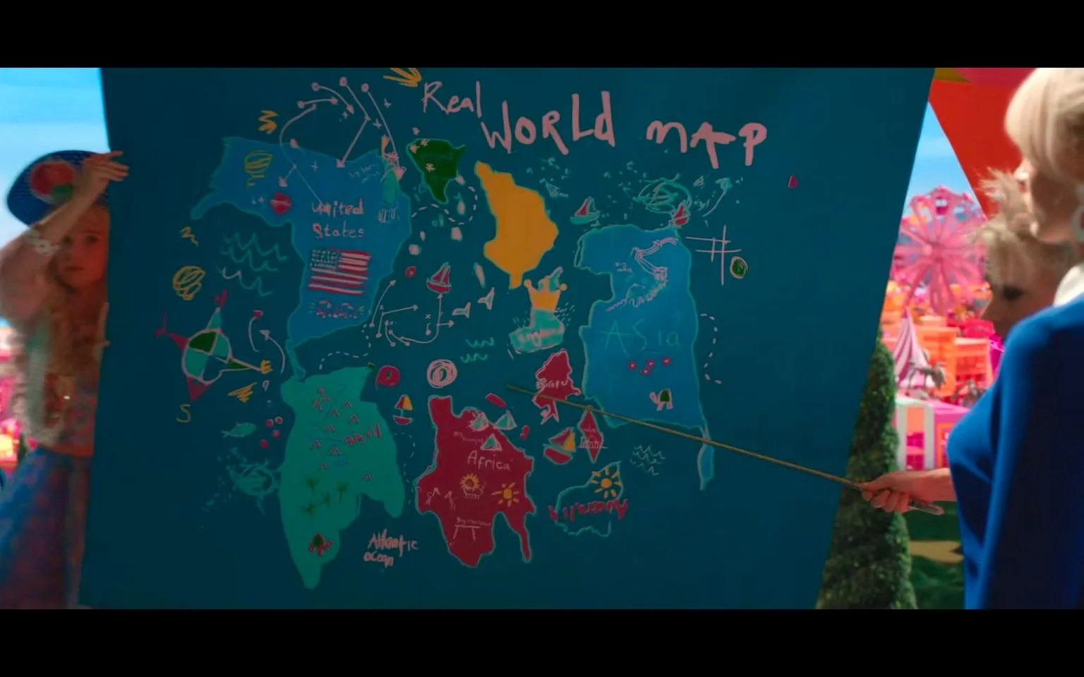 The image depicts a colorful and whimsical map labeled as the "Real World Map" from the movie "Barbie." The map features exaggerated and humorously inaccurate representations of continents and countries. The United States is prominently labeled and includes a small American flag. Other regions such as Asia and Africa are also marked, along with the "Atlantic Ocean." The map is hand-drawn with bright colors and playful illustrations, making it look more like a child's art project than a realistic world map.
In the scene, a person holds a pointer, directing attention to various parts of the map. The setting appears to be outdoors with vibrant, fantasy-like elements in the background, including a Ferris wheel and colorful structures. The atmosphere is playful and imaginative, fitting the whimsical theme of the "Barbie" universe. The style is cartoonish, with a focus on bright colors and exaggerated shapes.