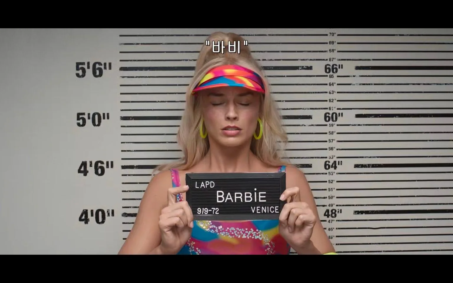 ```
The image depicts a character named Barbie in a mugshot-style setting. Barbie is standing in front of a height measurement chart, typically found in police stations for mugshots. Her eyes are closed, and she is holding a black nameplate with "LAPD", "BARBIE", and "VENICE" written on it, suggesting the setting is a fictional arrest scene.
Barbie is wearing a vibrant, multicolored visor and matching top, with large yellow hoop earrings, emphasizing her colorful and fashionable style. Her long blonde hair flows down her shoulders. The background is a standard height chart with measurements in feet and inches, indicating her height. The image has a comic or theatrical mood, playing into the playful and iconic nature of the Barbie character. The Korean subtitle "바비" translates to "Barbie". The lighting is bright, highlighting the vivid colors of her outfit and accessories.