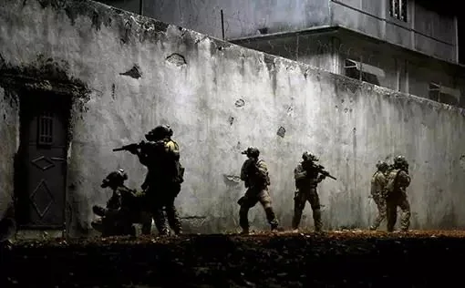 The image depicts a tactical operation scene at night, showing a group of six soldiers dressed in military gear. They are positioned against a weathered, textured wall, possibly part of a building, suggesting an urban environment. The soldiers wear helmets and tactical vests, equipped with firearms, indicating a high-alert situation. Their postures are alert and strategic, suggesting a coordinated entry or surveillance operation. The lighting is dim, with shadows cast dramatically on the wall, adding to the tense and secretive atmosphere. The color palette is muted, dominated by shades of gray and black, enhancing the scene's stealthy and covert nature. The composition captures a moment of anticipation, with the soldiers ready for action, highlighting the themes of military precision and preparedness. No identifiable text or branding is visible in the image, focusing solely on the tactical activity. The overall mood is intense and suspenseful, characteristic of night-time military maneuvers.