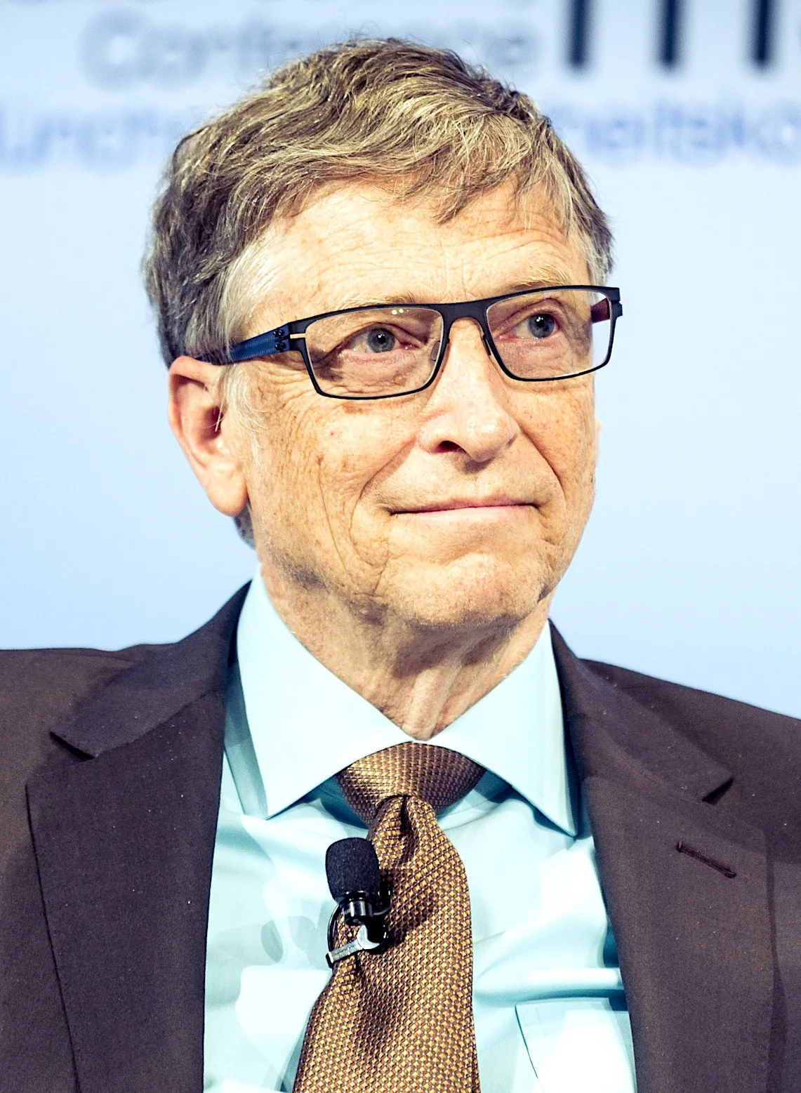 Bill Gates is shown in this image, captured in a professional setting, likely during a conference or interview. He is wearing a dark suit paired with a light blue shirt and a patterned brown tie, exuding a formal and polished appearance. Gates is seen with a calm and thoughtful expression, indicative of a moment of contemplation or attentive listening. His glasses add to his intellectual persona, and a microphone attached to his tie suggests he is either speaking to an audience or being interviewed. The background is blurred with indistinct text, but it contributes to the formal atmosphere, highlighting the context of a seminar or a discussion panel. The lighting is bright and even, focusing on Gates, enhancing the clarity and detail of his features. Overall, the image conveys a sense of authority and expertise, fitting for a prominent figure in the technology and business sectors.