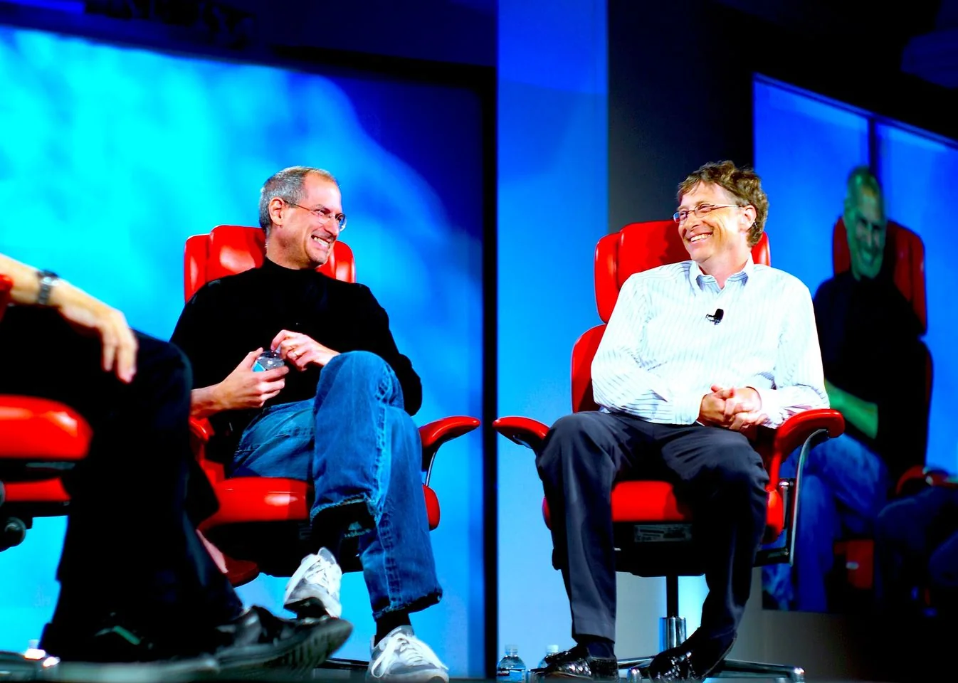 In this image, Steve Jobs and Bill Gates are seated on stage during what appears to be a conference or interview setting. They are both sitting in distinctive bright red chairs, which add a striking contrast to the blue background. Steve Jobs, on the left, is dressed in his signature black turtleneck sweater and blue jeans, a style he was well-known for. He has a relaxed demeanor, holding a small object in his hands, possibly a water bottle, and is smiling warmly. On the right, Bill Gates is wearing a light-colored, striped button-up shirt and dark pants. He is also smiling, and the two seem to be sharing a moment of mutual camaraderie. The lighting is bright, highlighting their expressions and features clearly. This image captures a significant moment between two iconic figures in the tech industry, known for their contributions to Apple and Microsoft, respectively. The backdrop is simple, with a screen reflecting their images, which adds depth to the scene.