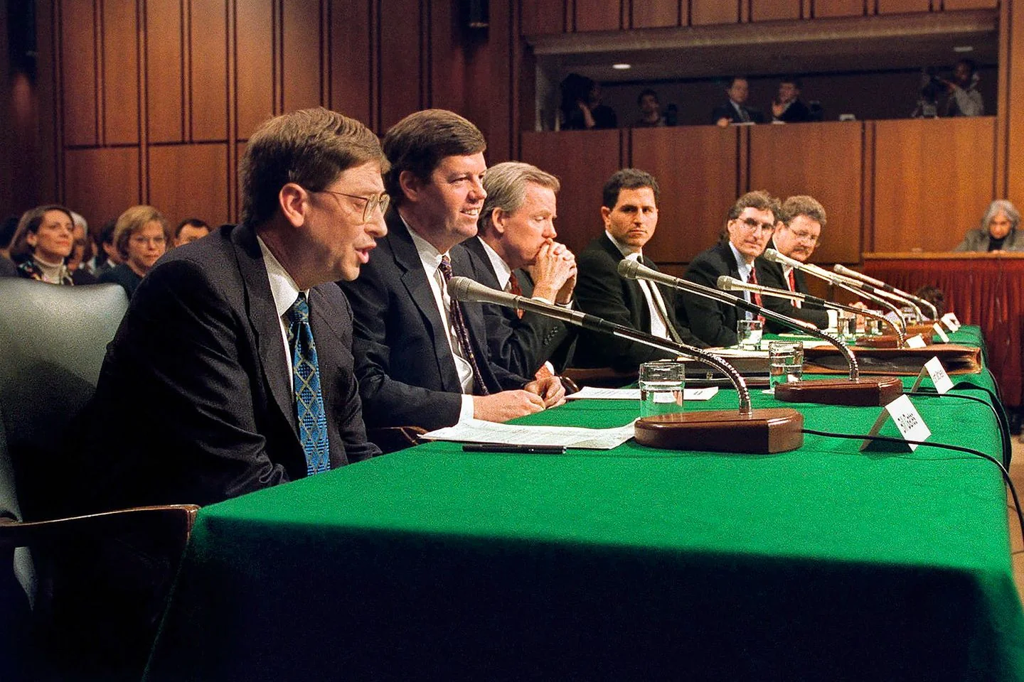 Bill Gates is depicted in this image, speaking at a panel or hearing. He is seated at a long table covered with a green cloth, alongside several other individuals. Each person has a microphone in front of them, suggesting a formal setting such as a congressional hearing or a similar event. Gates, in particular, is wearing a dark suit with a white shirt and a patterned tie, appearing focused on delivering his testimony. The room is large with wood-paneled walls, and several people are seated in the background, some observing the proceedings. The atmosphere is serious and professional, indicative of a high-stakes discussion or inquiry. There are nameplates and glasses of water on the table, emphasizing the formal nature of the event. The lighting is bright, typical of an indoor conference setting.