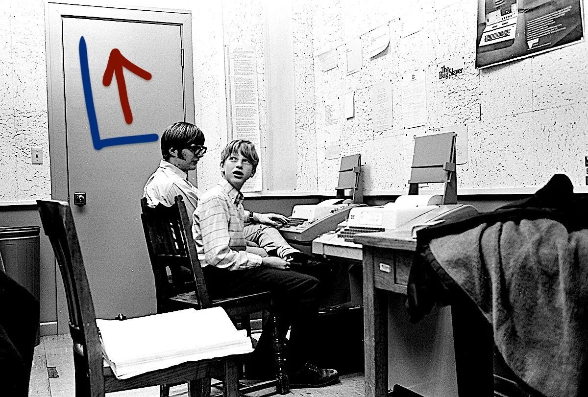 The black and white image captures a young Bill Gates, suggested by the hint, seated in an office setting. He is accompanied by another individual, both focused on computer terminals. The room is equipped with vintage computing equipment, indicative of the early era of technology. The walls are adorned with various papers and a visible poster titled "The Bug Slayer," suggesting a tech-focused environment. Both individuals are casually dressed, wearing button-up shirts. The setting is a simple office, featuring wooden chairs and a desk cluttered with papers. The mood conveyed is one of concentration and collaboration, likely during an early phase of computer innovation. The lighting is soft, typical of an indoor work environment, and the overall composition emphasizes a moment of historical significance in the tech world.