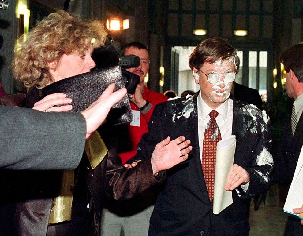 Bill Gates is featured in this image, captured during an unexpected event where he appears to have been hit with a pie. He is wearing a formal suit with a white shirt and a patterned tie, and holds some documents in his left hand. The pie has splattered across his face and suit, creating a surprising and somewhat humorous scene. The setting appears to be indoors, possibly in a public building or conference space, with multiple people surrounding him. A woman in the foreground is holding a black purse, and there are several onlookers, including a person with a camera, suggesting a media presence. The lighting is bright and even, typical of indoor public events. The atmosphere is chaotic, as people react to the incident. There is no visible text in the image to provide further context.