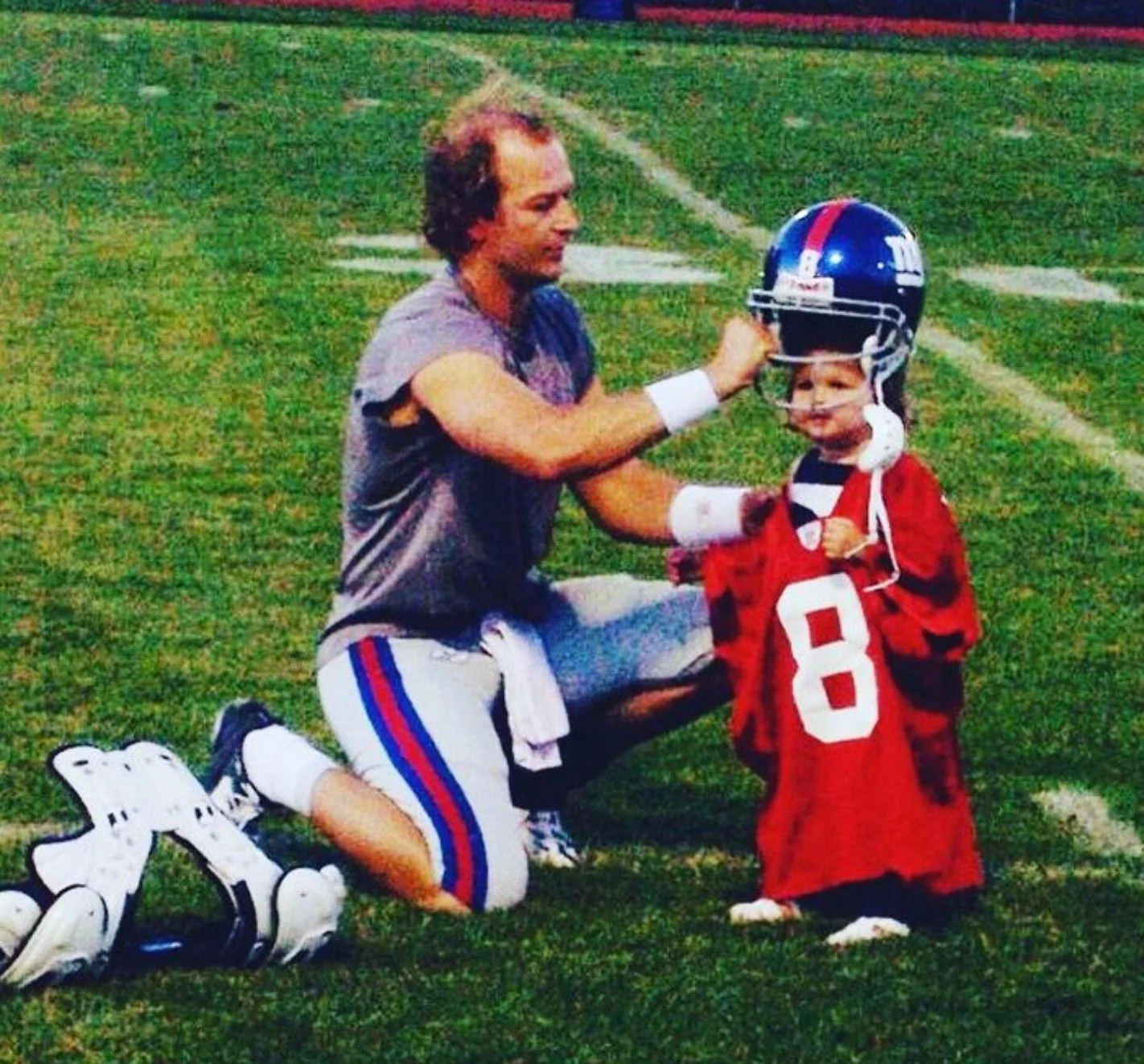 The image captures Tim Hasselbeck, a well-known figure associated with football, interacting on a football field. He is kneeling on the grass, assisting a young child who is wearing an oversized football helmet and jersey. The child’s jersey is bright red with the number 8 prominently displayed, almost engulfing their small frame. Tim Hasselbeck is dressed casually in athletic gear, consisting of a sleeveless gray top and shorts with blue and red stripes. His expression is kind and attentive as he adjusts the helmet on the child, creating a warm and engaging scene. The setting is a typical football field with visible yard lines and a distant running track in the background, suggesting this is a casual moment possibly after practice or a game. The lighting is natural, indicating it’s likely daytime, and the overall mood of the scene is heartwarming and familial.