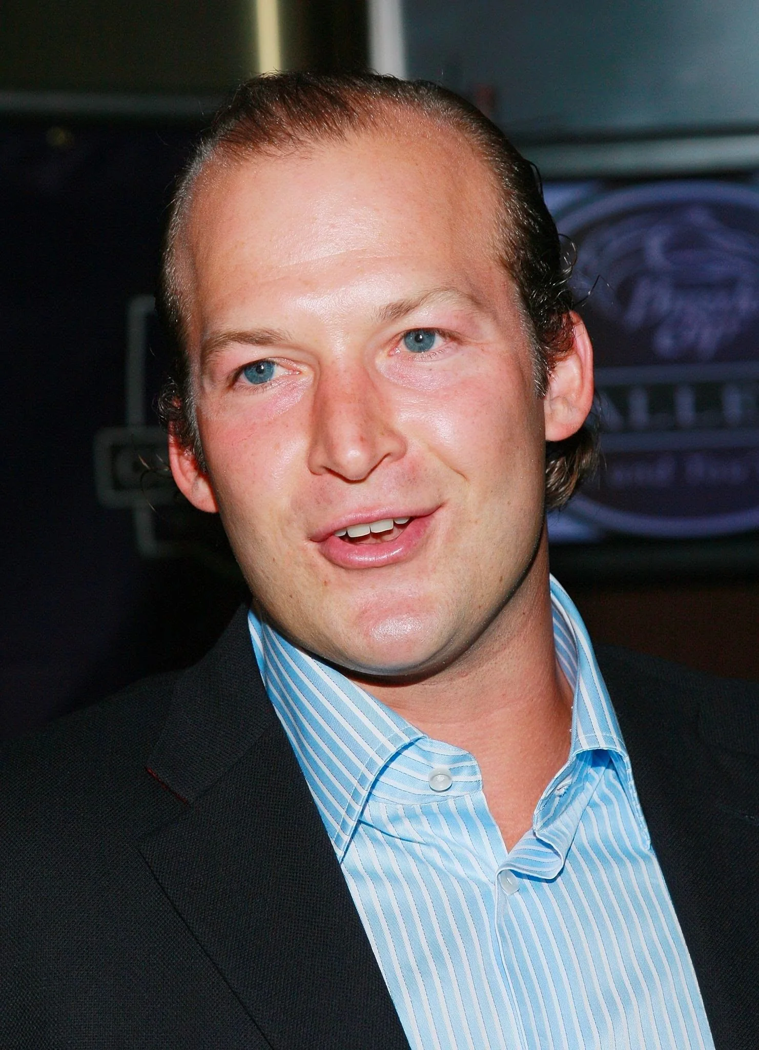 The image features Tim Hasselbeck in an indoor setting, likely at an event or gathering. He is dressed in a formal outfit, wearing a dark blazer over a light blue, striped dress shirt. The lighting is bright, highlighting his face and attire, suggesting this is a professional photograph taken at a media event or similar occasion. The background is softly blurred but hints at some event signage or branding, adding context to the setting.
Tim Hasselbeck's expression is relaxed, and he appears to be mid-conversation or speaking to someone. His hair is neatly styled, and he exudes a confident and approachable presence. The composition focuses on his face and shoulders, typical of a portrait style used during public appearances or interviews. The overall mood is professional and engaging, capturing a moment of interaction typical of public events.