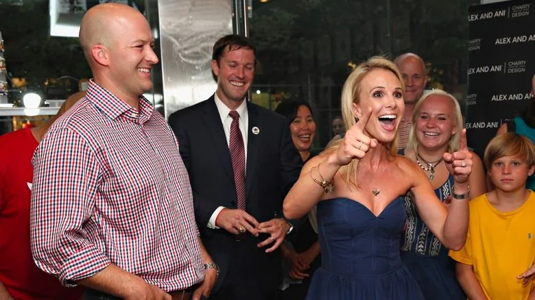 The image shows an indoor event with a festive and lively atmosphere. In the center, Elisabeth Hasselbeck is expressing excitement, gesturing with both hands and smiling broadly. She is wearing a strapless dark blue dress and a heart-shaped necklace, exuding a joyful energy. To her left, Tim Hasselbeck is present, dressed casually in a red and blue checkered shirt, looking towards Elisabeth with a smile, contributing to the cheerful mood.
Behind them, several other people are visible, including a man in a suit and tie, who seems to be laughing, and others who are smiling or looking towards the couple. The overall scene suggests a celebration or special occasion. The background is adorned with branding text "ALEX AND ANI" and "CHARITY DESIGN," which indicates the possible sponsorship or theme related to charity. The lighting is bright, emphasizing the festive ambiance of the gathering.