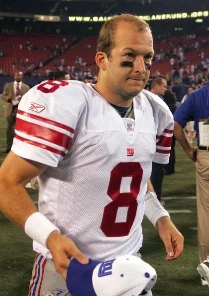 In this image, a football player is seen wearing a white jersey with the number 8 prominently displayed in red on both the front and sleeve. The jersey also features the logo of the New York Giants, indicating that this is an NFL player. He has black eye paint under his eyes, a common practice among football players to reduce glare from stadium lights. The player is holding a cap in his left hand and appears to be walking on the field, likely after a game. The background shows a stadium setting with a crowd visible in the stands and other people on the field, suggesting a post-game scene. The lighting in the image is bright, illuminating the player and highlighting his jersey’s details. The overall mood captures a moment of transition, possibly as the player exits the field.