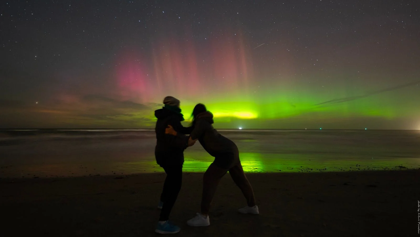 The image captures a stunning nighttime scene on a beach, with two silhouetted individuals posing playfully in the foreground. The sky is alive with vibrant displays of green and pink auroras, casting a magical glow over the scene. The individuals are bundled up in winter clothing, suggesting a chilly atmosphere, and their playful interaction adds a sense of joy and wonder to the composition.
The auroras stretch across the sky, with streaks of green dominating the center and soft pink hues to the side, reflecting gently on the water. Stars are visible against the dark sky, enhancing the ethereal quality of the setting. The beach is calm, with gentle waves lapping at the shore, and the horizon is illuminated by a distant light, possibly from a lighthouse or a ship. The overall mood is serene and awe-inspiring, capturing the natural beauty of the aurora borealis.
- Key OCR lines: "Saturday, September 23rd."