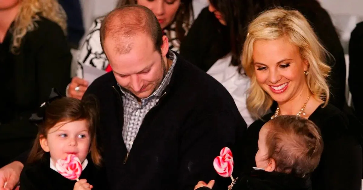 Tim and Elisabeth Hasselbeck are pictured in this warm and family-oriented setting. The image captures a candid moment where Tim, wearing a dark sweater over a checked shirt, is looking down, appearing engaged and attentive. Elisabeth is seated to his right, smiling warmly at a child in her arms. She is elegantly dressed in a black outfit and accessorized with a gold necklace, her blonde hair styled casually. The child in Elisabeth's arms is holding a red and white swirled lollipop, as is another young girl sitting beside Tim, who looks towards the camera. The background shows other seated individuals, suggesting this might be at a public event or gathering. The atmosphere is cozy and familial, with the focus on shared moments and interactions among the family members. The lighting is soft, highlighting the warmth and joy in their expressions.