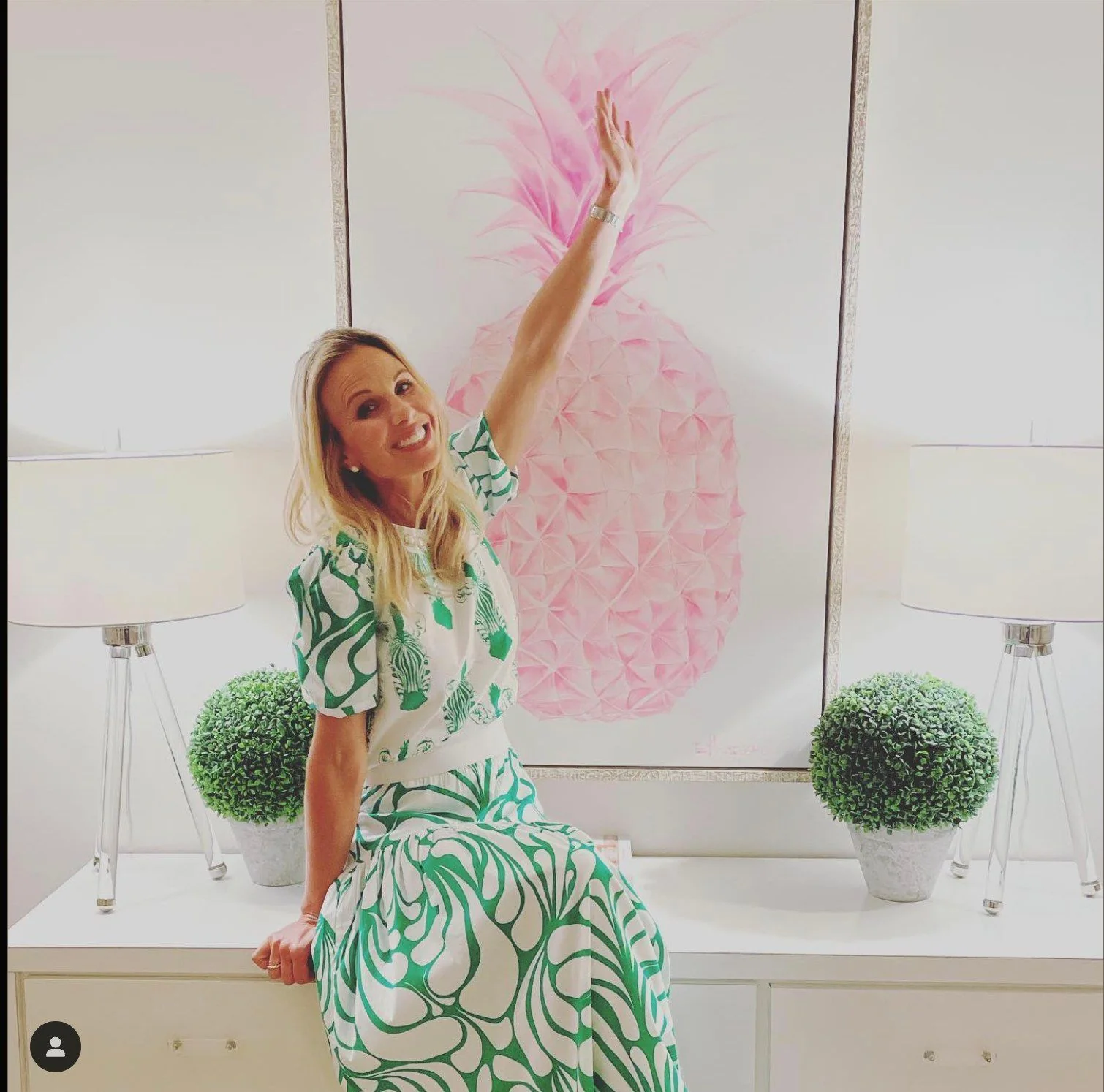 Elisabeth Hasselbeck is shown sitting on a white cabinet in a stylish indoor setting. She is wearing a matching outfit featuring a bold green and white pattern with swirls. Her top is short-sleeved, and she pairs it with a long, flowing skirt. The expression on her face is joyful and welcoming, as she raises one arm upwards. Behind her, there's an eye-catching artwork of a large pink pineapple, which adds a vibrant pop of color to the background. The setting is enhanced by two identical lamps on either side, with green spherical plants in pots next to them, giving the room a balanced and aesthetically pleasing appearance. The lighting is bright and warm, creating an inviting atmosphere. The composition highlights both Elisabeth and the artistic elements of the room, blending fashion and decor seamlessly.