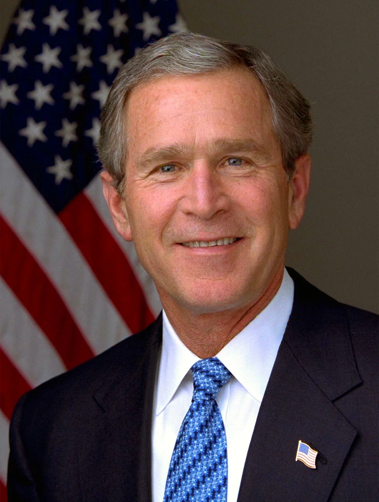 The image is a formal portrait of President George W. Bush. He is positioned in front of a blurred American flag, suggesting a patriotic and official setting. The composition is typical of a presidential portrait, with President Bush centered in the frame, conveying a sense of authority and presence. He is wearing a dark suit jacket paired with a white dress shirt and a blue patterned tie. A small American flag pin is visible on the lapel of his jacket, reinforcing the patriotic theme. The lighting is even and bright, highlighting his facial features and giving the portrait a polished and professional appearance. There is a calm and composed expression on his face, typical of official portraits, aiming to convey confidence and leadership. This image likely served as an official representation during his time in office.
