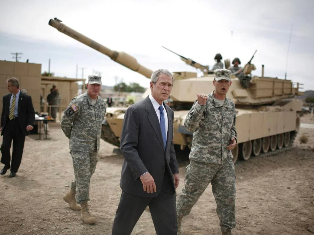 President George W. Bush is shown in this outdoor scene, set against the backdrop of a military base. The image captures him walking on a dirt path, dressed in a formal dark suit with a light blue tie. His expression appears focused and serious. To his right, a military officer in a camouflage uniform and cap is engaged in conversation, gesturing with his hand, possibly explaining something. In the background, another service member stands at ease. A large tan military tank is prominently displayed, with two soldiers visible on top, equipped with helmets. The setting conveys a sense of authority and military operations, with the tank adding to the visual prominence of the scene. The lighting is natural, suggesting a clear day. The composition highlights a moment of interaction between the president and military personnel, emphasizing themes of leadership and defense.