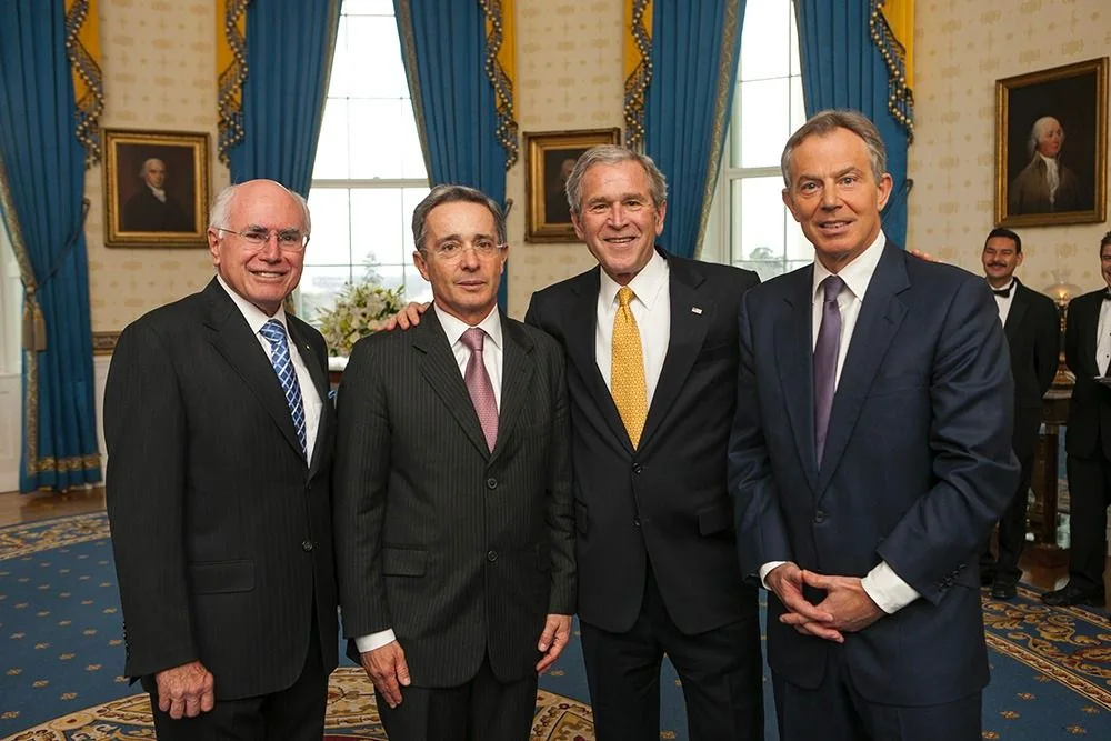 The image shows a formal gathering of four men in a well-decorated room with blue curtains and portraits on the walls, suggestive of an official setting. The individuals are dressed in suits, indicating a formal or diplomatic event. According to the hint, one of the men is labeled as "GW Bush," smiling and positioned centrally. To his right, another man stands with a similar demeanor, and the hint identifies this person as "Tony Blair." On the left, another individual is present, and the hint mentions "Dick Cheney." The room's decor, including the ornate carpeting and classical portraits, adds to the formality of the scene. The lighting is bright, emphasizing the distinguished and cordial atmosphere of the meeting.