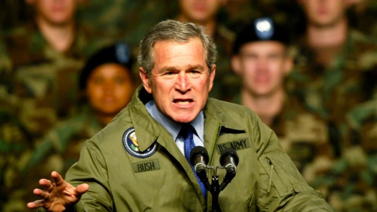 President George W. Bush is delivering a speech at a military event. He is prominently positioned at a podium with two microphones, wearing a green jacket featuring U.S. Army insignia and his name, "BUSH," embroidered on the front. The presidential seal is visible on the jacket, emphasizing the official nature of the setting. In the background, a group of military personnel listens attentively, dressed in camouflage uniforms and berets, highlighting the context of the event. Bush's expression is serious and determined, possibly conveying the gravity of his message. The lighting is bright, focusing on him, while the background remains slightly out of focus, directing attention to the president. This setting likely represents an important announcement or address to the troops.