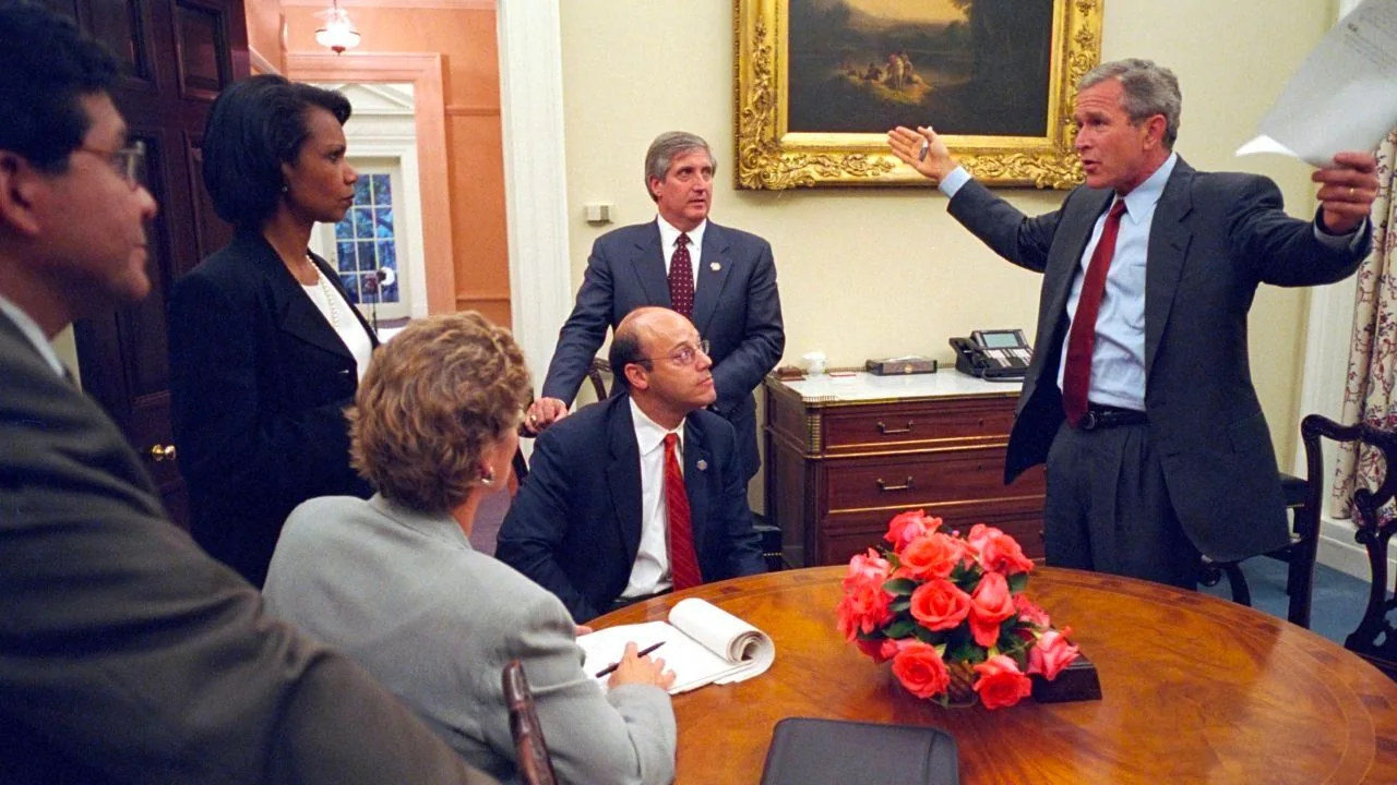 The image depicts a meeting in what appears to be a formal office setting. The room has warm lighting and features elegant furniture, including a round wooden table adorned with a vase of pink roses. In the background, a large, ornate painting hangs on the wall above a wooden sideboard. To the right, a man in a suit, referred to as "Bush" in the hint, is gesturing with his arms spread wide, holding some papers in his hand. He appears to be speaking passionately, conveying a sense of urgency or importance.
Seated around the table, several individuals listen attentively. One of them, identified as "Condi," stands out as she is standing and seems engaged in the discussion. She is wearing a dark blazer and a white blouse. The others, seated and standing, are dressed in formal attire, indicating the seriousness of the meeting. Their expressions vary from attentive to contemplative. The setting and the demeanor of the participants suggest a high-level political meeting.