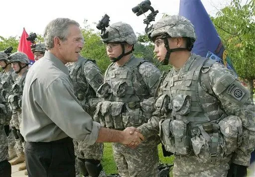 President George W. Bush is seen outdoors shaking hands with a soldier in military uniform. The setting appears to be a daytime event, likely a formal military gathering or visit. The soldiers are dressed in full combat gear, complete with camouflage uniforms, tactical vests, and helmets equipped with night-vision goggles. The soldier shaking hands with President Bush is positioned to the right of the image and is engaged in a formal handshake, looking directly at the president. Other soldiers stand in line, also in full gear, with attentive postures. President Bush is dressed in civilian clothing, wearing a light gray button-down shirt and dark trousers. The mood is formal and respectful, indicative of a ceremonial or official military event. In the background, greenery and flags are visible, contributing to the formal and organized atmosphere of the scene. The composition captures a moment of interaction between military personnel and the president, highlighting themes of leadership and respect.