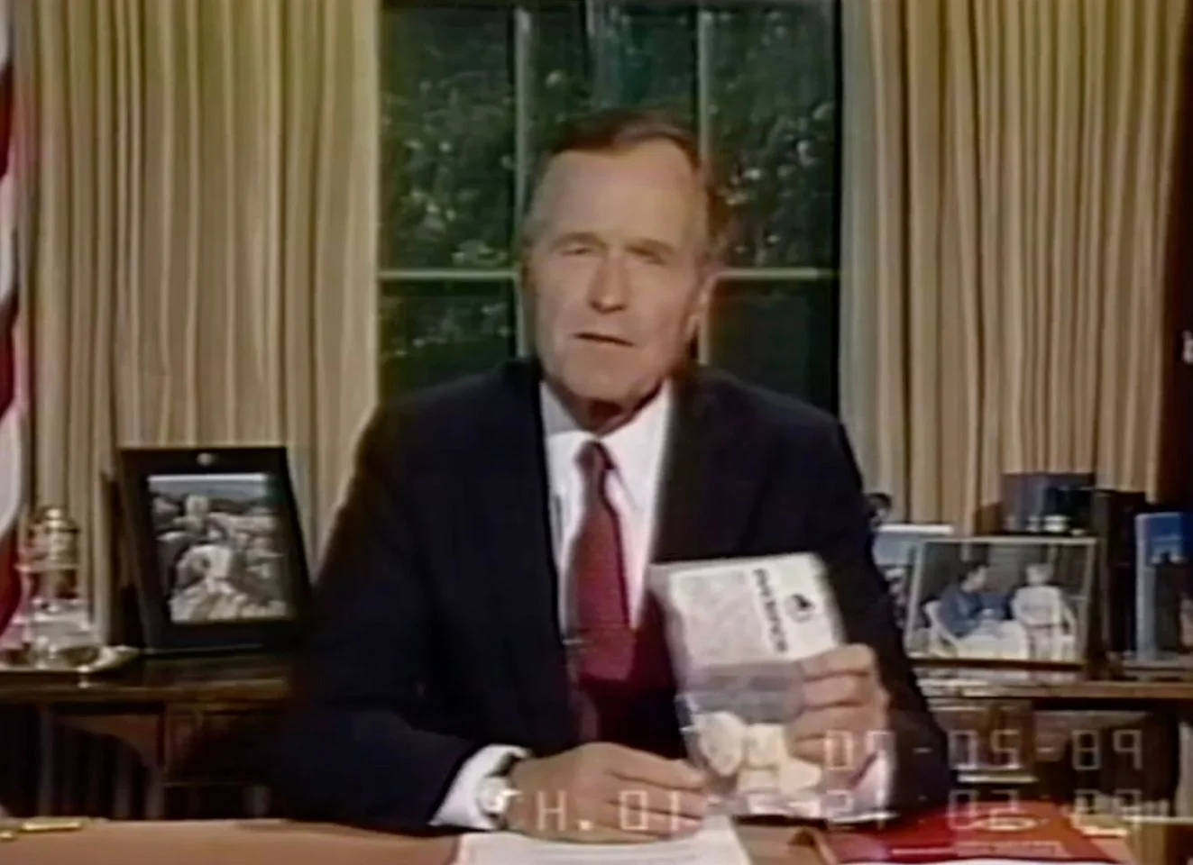 1) Rich description: The image shows a video frame of HW Bush, seated at a desk in a formal setting, likely the Oval Office, given the recognizable backdrop of curtains and a large window. There are family photographs on the desk, along with an American flag partially visible to the left. HW Bush is dressed in a dark suit with a white shirt and a red tie, conveying a sense of formality and authority. His expression appears serious as he holds a plastic bag containing an item, possibly a prop used to emphasize a point during a speech. The lighting is warm, highlighting the subject and the desk. The timestamp at the bottom of the screen suggests the footage is from July 5, 1989, which aligns with a period during his presidency when he addressed issues like drug policy. The presence of personal photographs adds a personal touch to the otherwise official setting.
2) On-screen text block:
- Key OCR Lines: "07-05-89", "CH. 01 21 05:09"