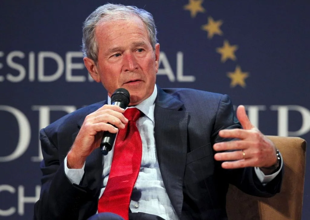 The image depicts President George W. Bush holding a microphone during what appears to be an event or discussion. He is seated indoors against a backdrop that includes partially visible text, notably the words "PRESIDENTIAL" and "CHILDREN HOSPITAL." President Bush is wearing a dark suit with a white shirt and a bright red tie, suggesting a formal occasion. His expression is thoughtful as he gestures with his left hand, indicating he might be emphasizing a point or responding to a question. The background is slightly blurred, focusing attention on him. The lighting is even and suitable for an indoor setting, likely a conference or public speaking venue. His posture is relaxed yet engaged, typical of someone participating in a conversation or interview. The setting and attire suggest this is a professional event, possibly related to a hospital or healthcare initiative.