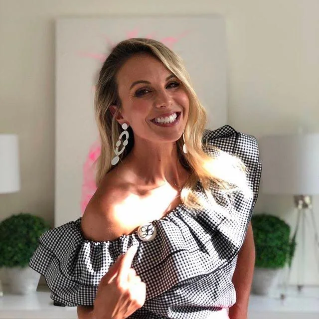 The image features a woman indoors, posing with a bright, cheerful smile. She wears a stylish off-the-shoulder top with a black and white checkered pattern and a large ruffle. Her long, wavy blonde hair cascades over her shoulders, and she accessorizes with eye-catching, dangling white earrings. The background is softly lit, suggesting a warm, welcoming environment. There are two lamps with white shades and green potted plants, adding a touch of elegance and freshness to the setting. The lighting is natural, creating a soft glow on her skin, enhancing the overall vibrant and joyful mood of the image. The composition focuses on her expressive, happy demeanor, highlighting both her fashion sense and the pleasant ambiance of the room. The overall atmosphere is lively and positive.
- Captions / subtitles: "You're the one that I wanna be with now"
