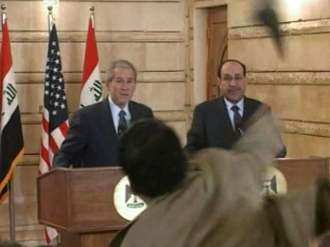 The image depicts a press conference scene featuring George W. Bush. He is standing at a podium alongside another individual, likely a political figure, in a formal setting with flags of Iraq and the United States prominently displayed in the background. Both individuals are dressed in formal suits, indicating a diplomatic or official event. The setting appears to be indoors, with a beige and wood-toned backdrop adding a formal atmosphere.
In the foreground, a shoe can be seen mid-air, being thrown towards George W. Bush. This moment captures a significant incident, reflecting a tense and unexpected interruption during the press conference. The expressions on the faces of those at the podium are focused, suggesting they are responding to or anticipating the unfolding event. The lighting is bright, ensuring clear visibility of the event and the individuals present. The scene is a still image from a video frame, capturing a moment of political drama and tension.