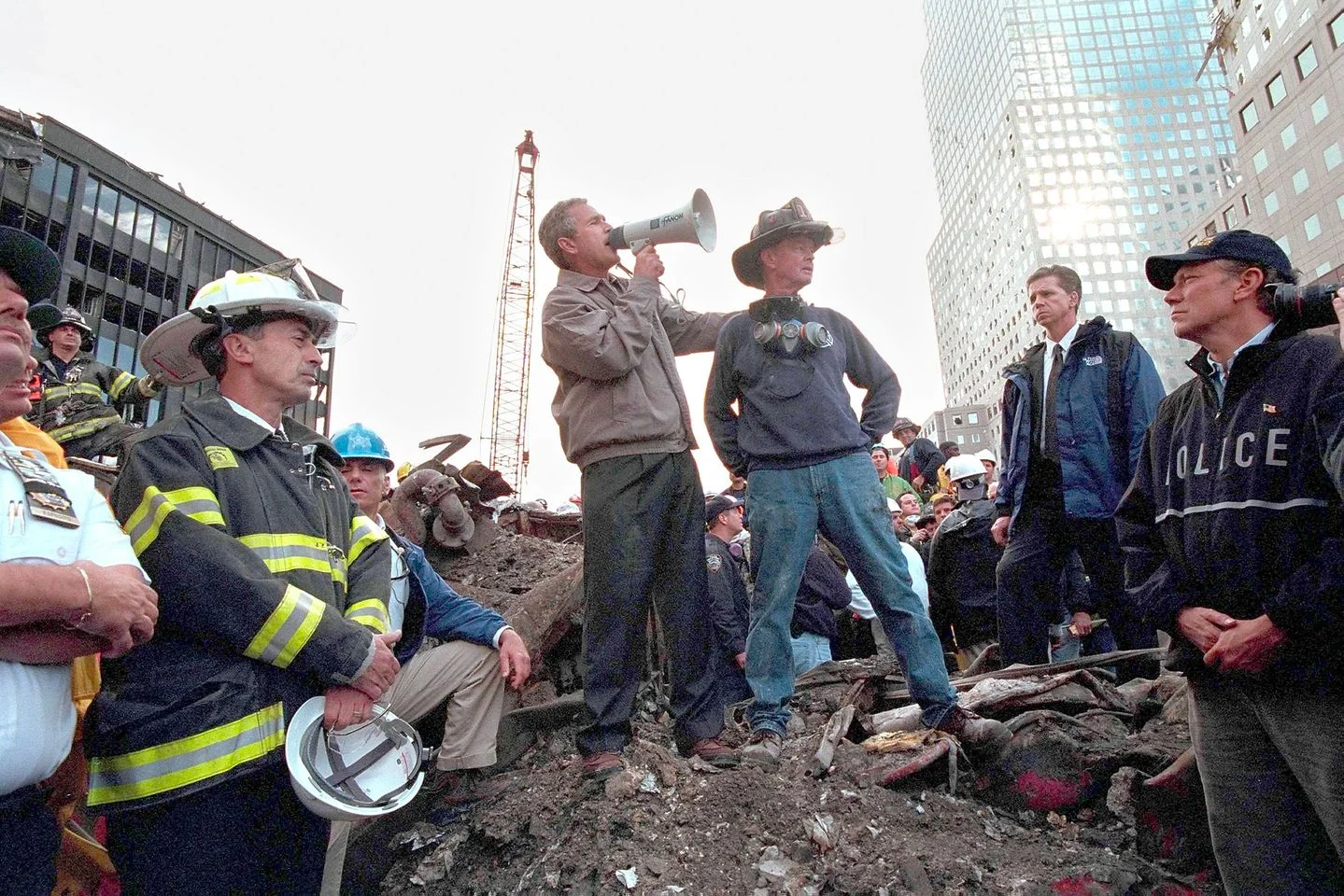 President George W. Bush is seen standing on a pile of rubble with a megaphone, addressing a crowd of emergency responders and other individuals. The setting is outdoors, amidst the wreckage of the 9/11 attacks. Surrounding him are firefighters and police officers, identifiable by their uniforms and safety helmets. The mood is solemn and determined, as the group listens to the president's words. Bush is positioned slightly elevated, giving him a commanding presence. The background includes damaged buildings and construction equipment, suggesting an ongoing rescue and recovery effort. The lighting is natural, coming from a clear sky, casting an even illumination over the scene. The image captures a moment of national resolve and unity in the face of tragedy.