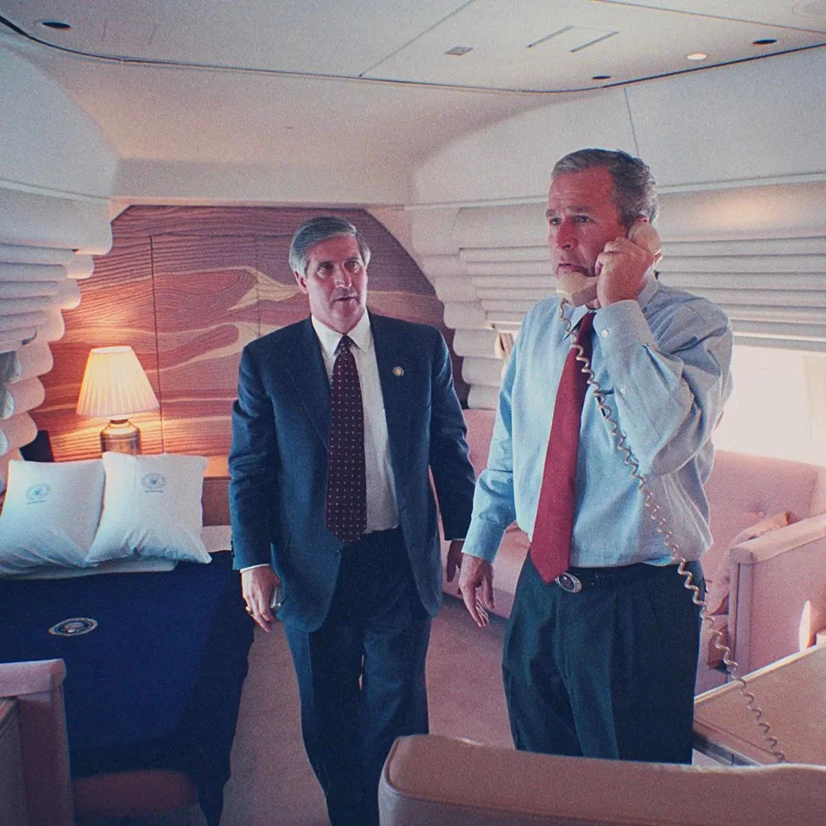The image shows President George W. Bush inside an aircraft, likely Air Force One, given the presidential seal visible on the bed pillow. He is holding a phone to his ear, appearing focused and possibly engaged in a serious conversation. President Bush is wearing a light blue dress shirt with the sleeves rolled up and a red tie, giving a sense of informality yet urgency. Beside him stands another man in a suit and tie, who appears to be attentively listening or waiting to assist. The setting is a well-appointed cabin with a lamp, plush seating, and a bed, suggesting a private area for rest and work. The lighting is soft, creating a calm yet professional atmosphere. The image captures a moment of leadership and communication within a high-level governmental context.