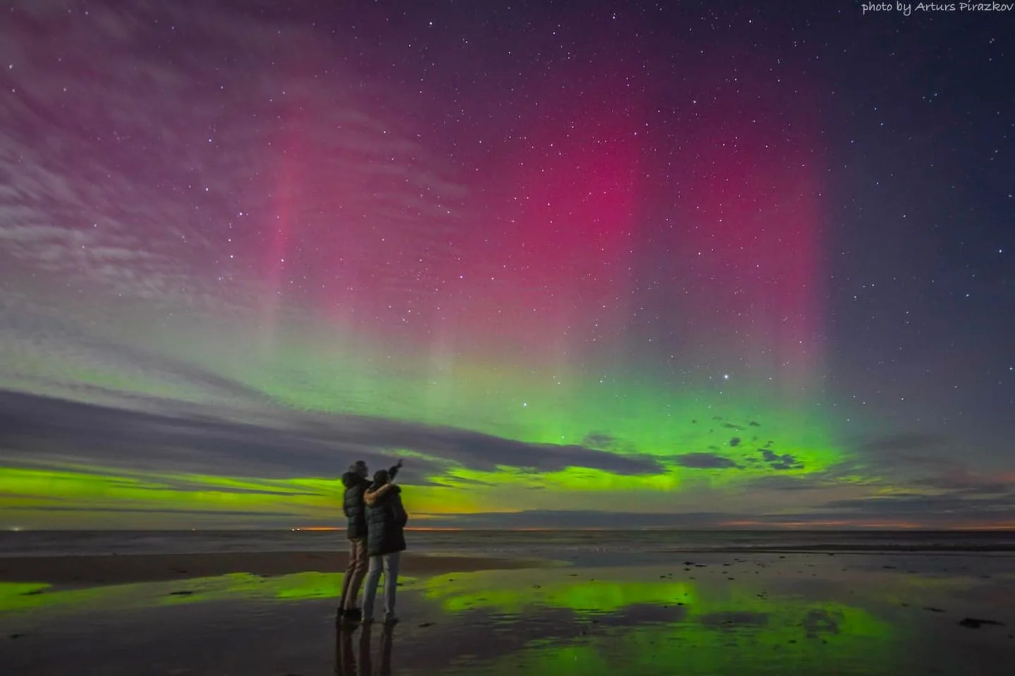 The image captures a breathtaking scene of the aurora borealis over a serene beach setting at night. In the foreground, a couple stands together, with one of them pointing towards the vibrant display in the sky. They are dressed warmly, suggesting a cool atmosphere, with coats that have fur-lined hoods. Their reflections are faintly visible on the wet sand beneath them, adding depth to the photograph.
The sky is a mesmerizing blend of colors, with vivid green and pink hues painting the heavens. The auroras create a curtain-like effect, rippling across the sky and punctuated by twinkling stars, which add to the ethereal quality of the scene. Light from the auroras also casts a gentle glow on the surroundings, creating a beautiful contrast with the darker beach and ocean.
In the top right corner of the image, the text "photo by Arturs Pirazkov" is visible, crediting the photographer for capturing this stunning natural phenomenon. The overall mood of the image is tranquil and awe-inspiring, as the couple seems to be absorbed in the beauty of the night sky.