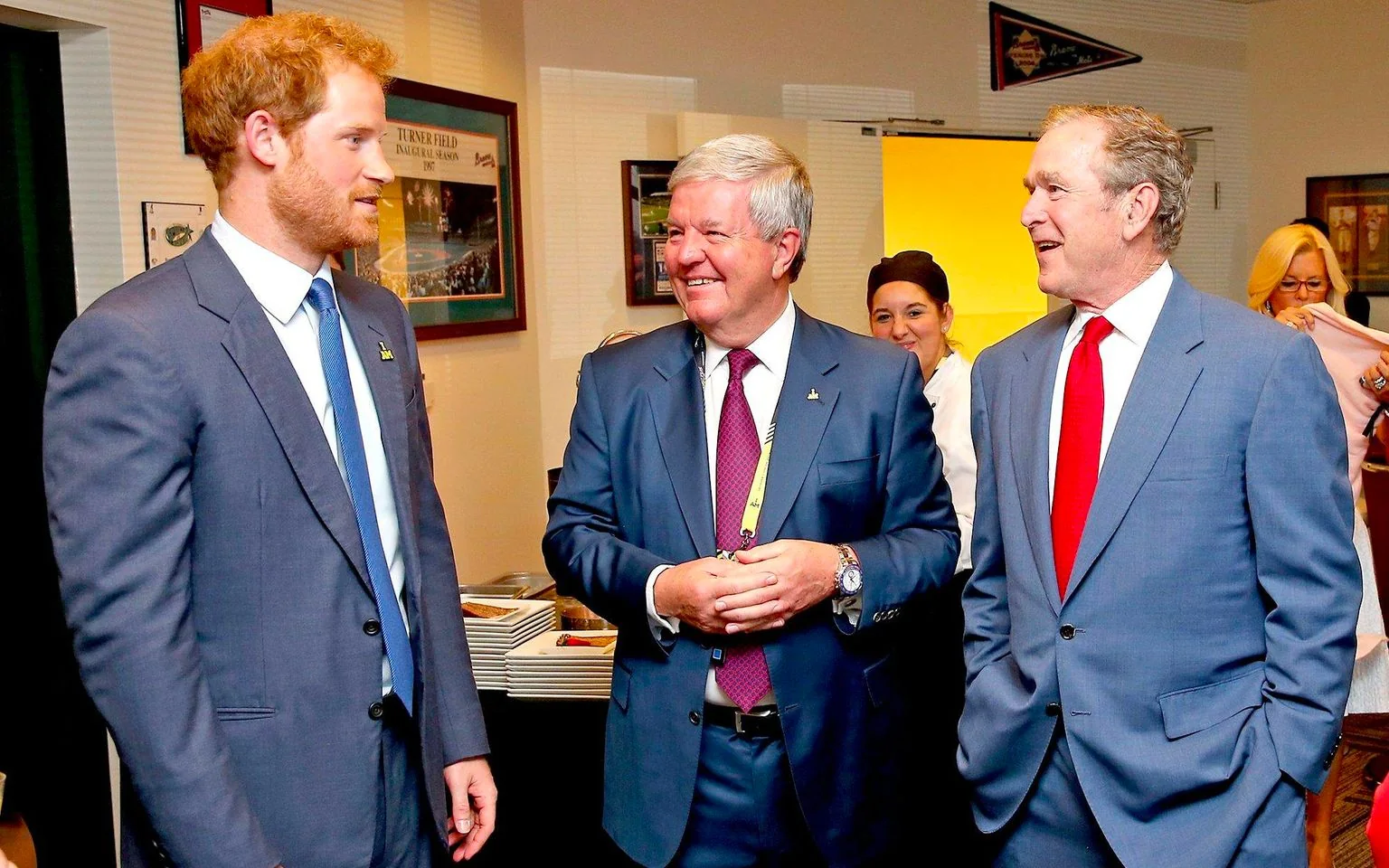 Prince Harry and President George W. Bush are pictured together in a warmly lit indoor setting. They are engaged in conversation with another man between them, all wearing formal suits. Prince Harry’s suit is a dark blue, paired with a light blue tie, while President Bush is also in a blue suit, accentuated with a striking red tie. The man between them is wearing a darker suit with a maroon tie and appears to be listening attentively, smiling as he engages with the two.
The environment has a professional and formal ambiance, possibly at an event or gathering, with framed pictures and memorabilia on the walls. Notably, there is a framed image with the text "TURNER FIELD INAUGURAL SEASON 1997," adding context to the location or event they are attending. In the background, there are stacks of plates and people who appear to be part of the catering staff, indicating a reception or social event setup. The overall mood is friendly and cordial, highlighting a moment of interaction between these notable figures.