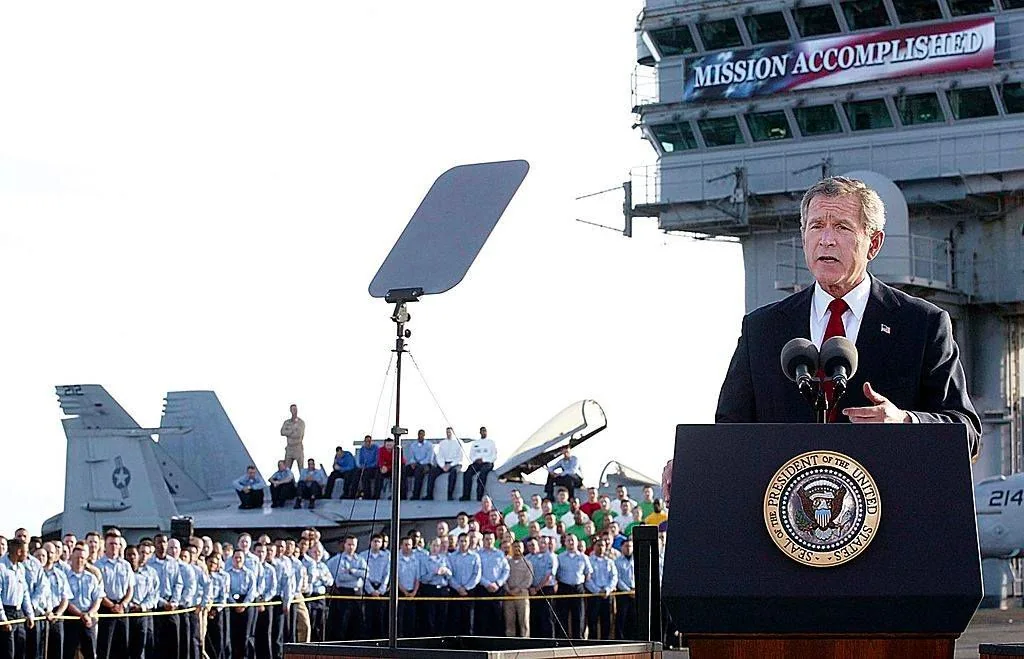 ```
The image captures President George W. Bush delivering a speech on the deck of a military ship. He is standing at a podium with the presidential seal prominently displayed. Behind him, a large banner with the words "MISSION ACCOMPLISHED" is visible, attached to the superstructure of the ship. The event takes place outdoors during the day, with clear visibility. In the background, military personnel in uniform are gathered, some standing in formation while others sit on aircraft parked on the deck. The scene conveys a formal and significant occasion, with the President dressed in a dark suit and red tie, speaking in front of microphones. The lighting is natural, indicating it is likely daytime, and the atmosphere appears serious yet triumphant, reflecting the message on the banner.